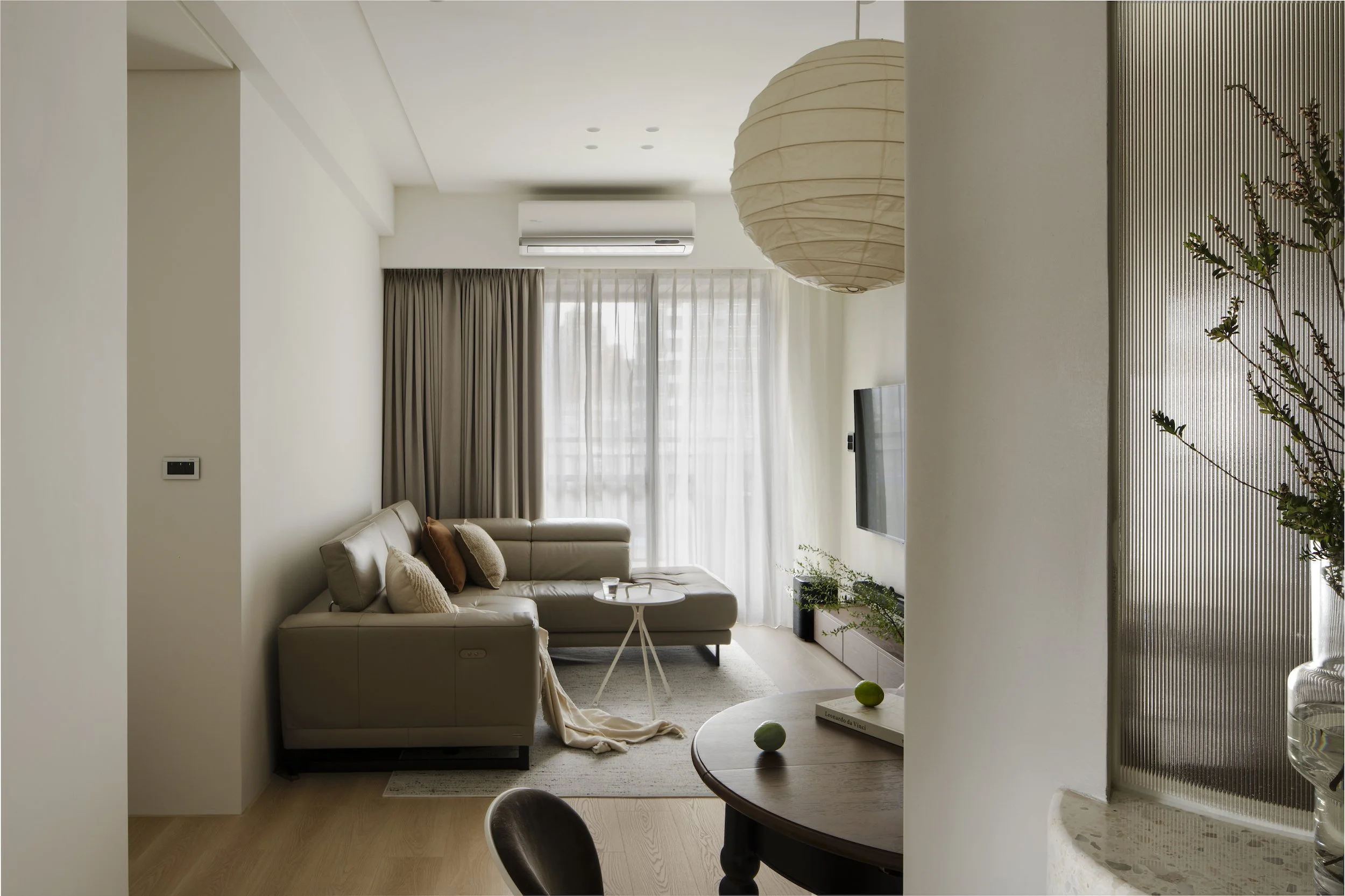Modern living room with beige sofa, small round white coffee table, and large window with sheer curtains. There are decorative pillows on the sofa, and a potted plant near the TV. A large paper lantern hangs from the ceiling.