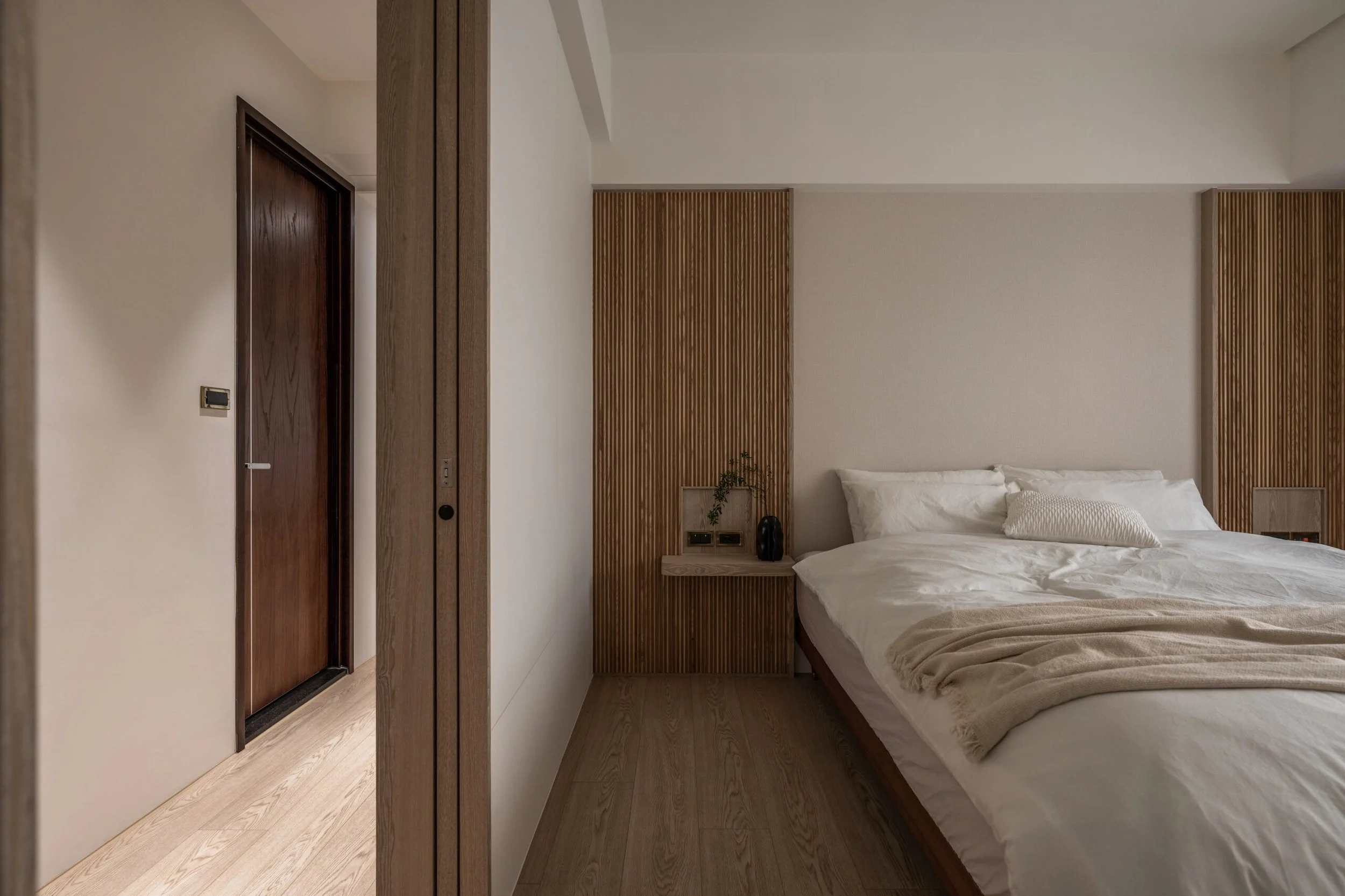A modern bedroom with a bed, wooden headboard, and a minimalist nightstand with a potted plant and black decorative item. White walls and wood flooring.