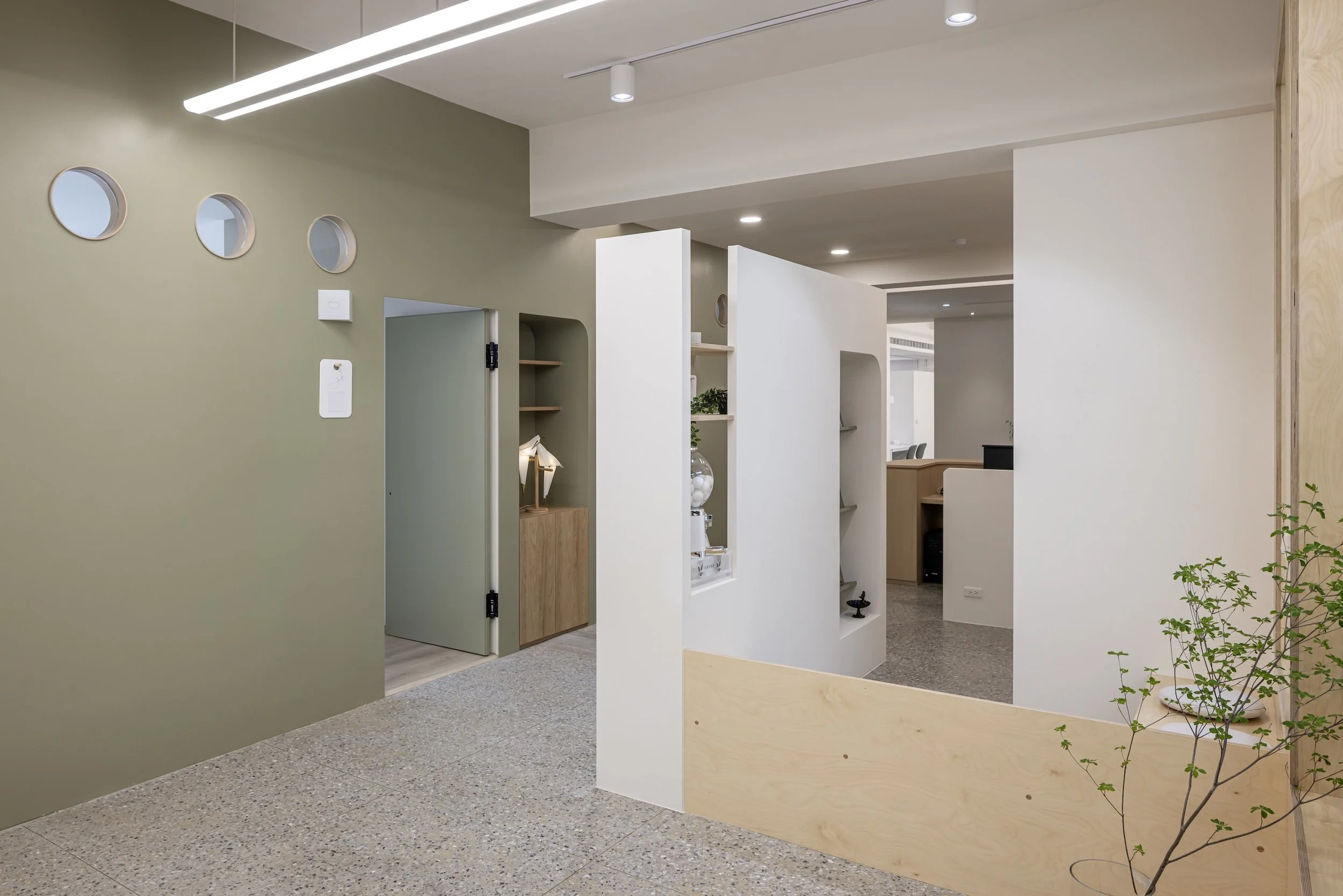 Modern office interior with beige and green walls, white partition walls, wooden accents, and potted plants.