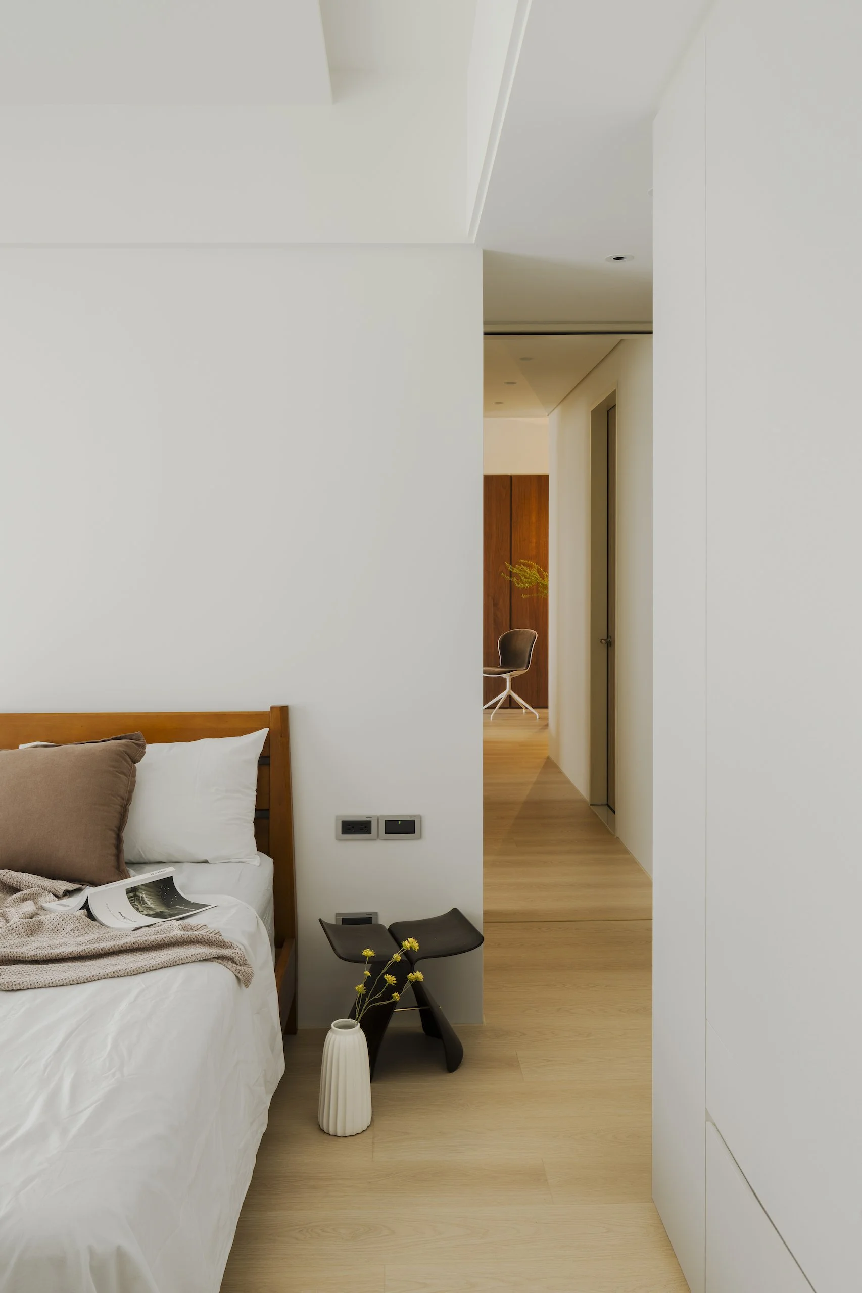 A bedroom with a bed, a small black side table with yellow flowers, a white vase, a brown pillow, an open book, and a beige blanket. A corner of the room reveals wooden flooring, a white wall with electrical outlets, a doorway leading to a hallway, and a wooden cabinet with a beige chair and yellow flower decoration in the background.