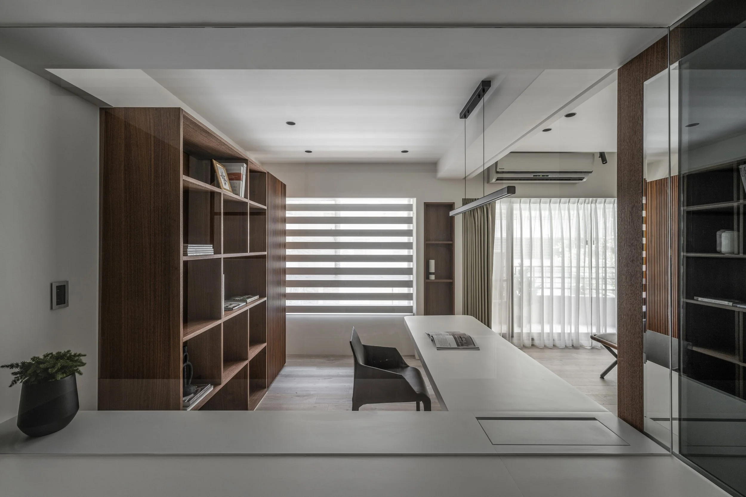 Modern interior living room with white walls and ceiling, wooden shelves, and large windows with horizontal blinds and curtains. There is a white table with chairs and decor items, including a black vase with a plant.