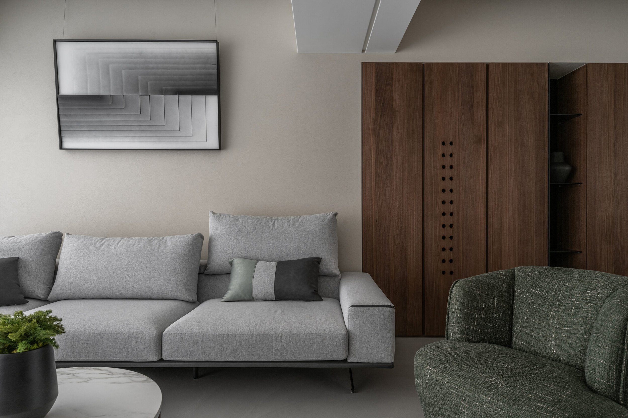 Modern living room with gray sofa and green armchair, black vase with green plant on white marble coffee table, abstract black-and-white wall art, wooden cabinet with small black holes and open shelves, beige wall.
