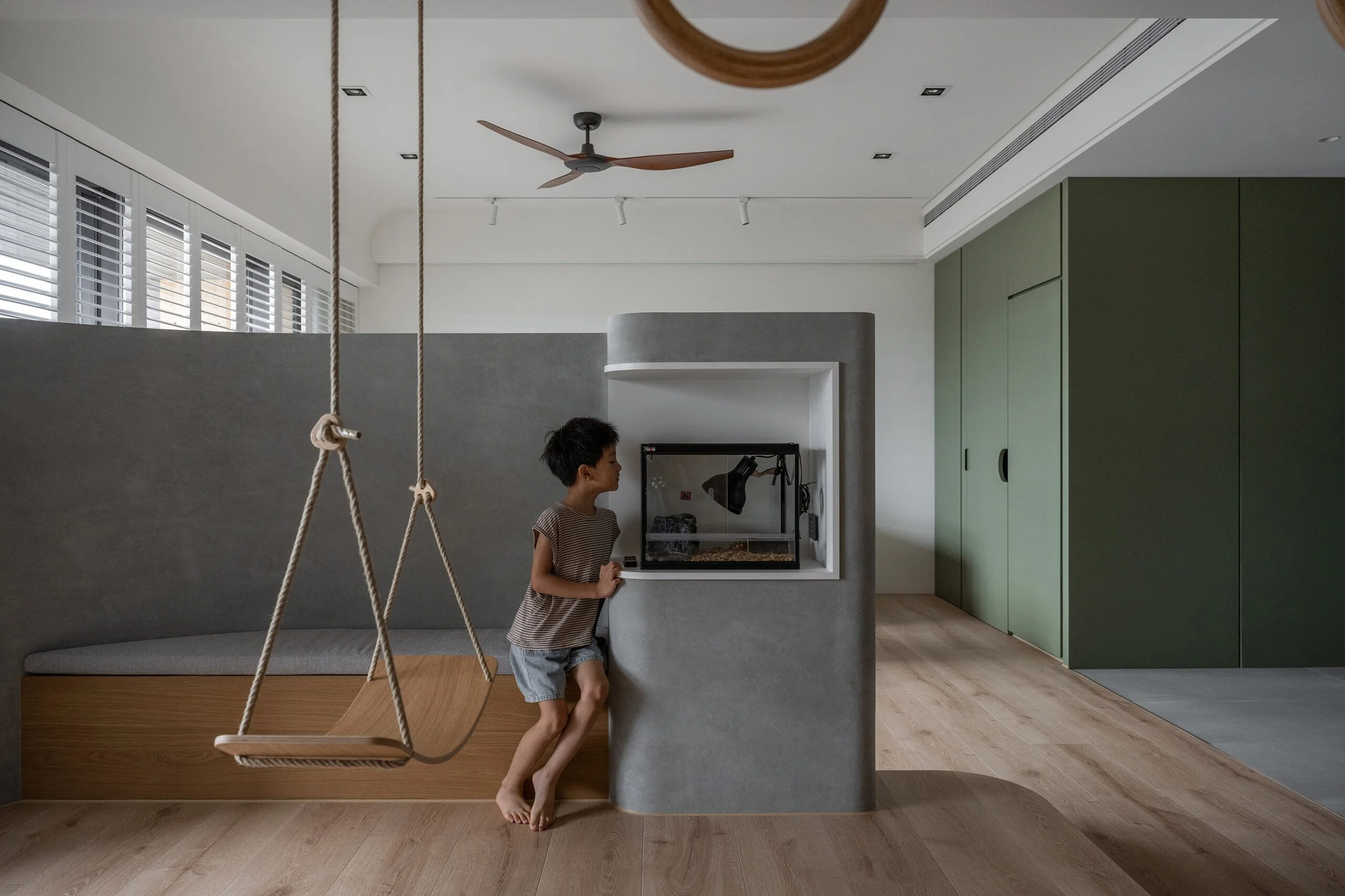 A young boy with short dark hair, wearing a striped t-shirt and shorts, stands barefoot by a modern, gray, curved partition in a spacious living room. Behind him is a swing made of wood and ropes hanging from the ceiling. The room has light-colored wooden flooring, green and white walls, a ceiling fan, and large windows with white blinds.