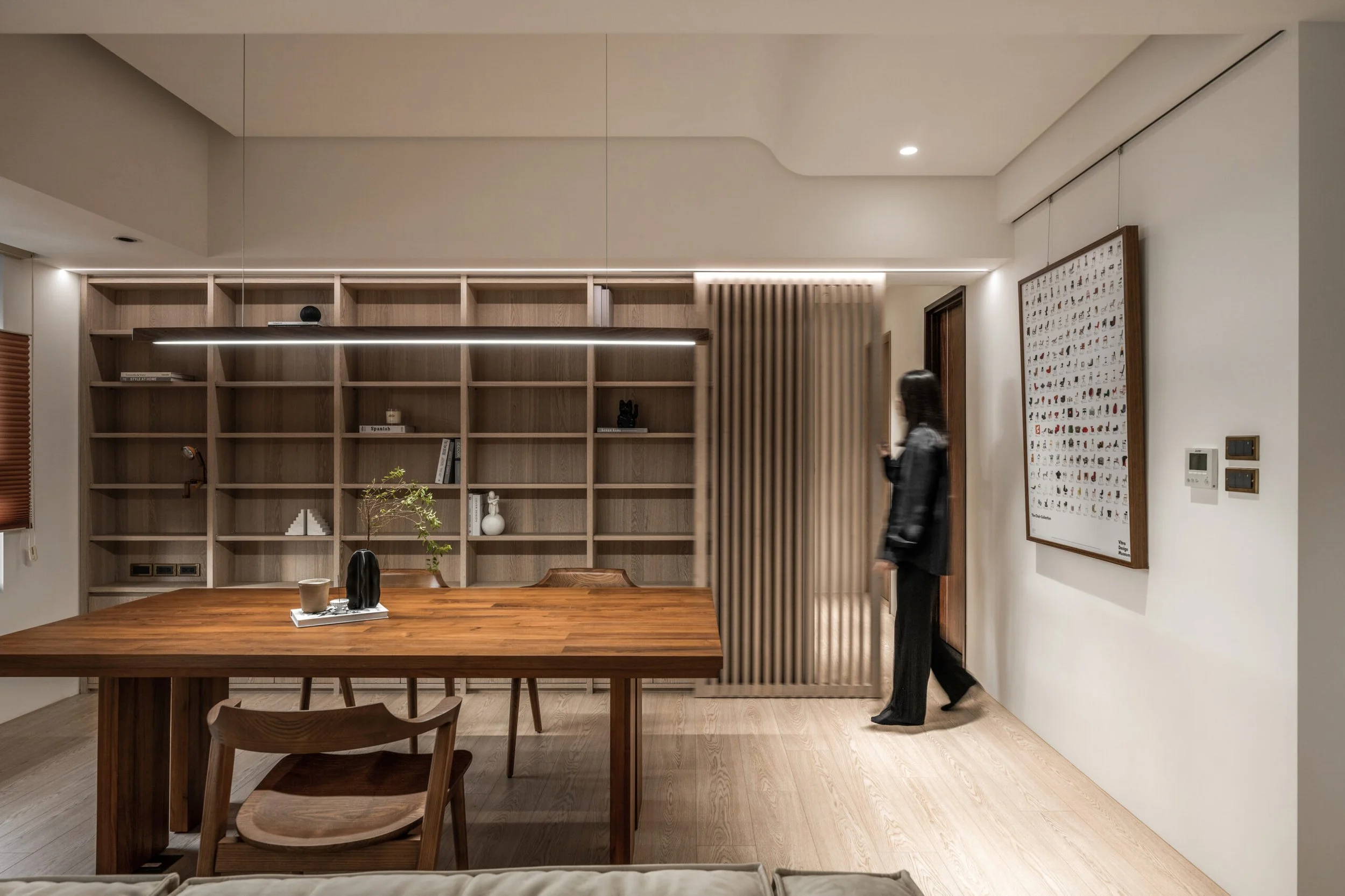 Modern living room with wooden bookshelf, a dining table with chairs, and a woman walking past a decorative slatted wooden partition, all illuminated by recessed lighting and a strip light.