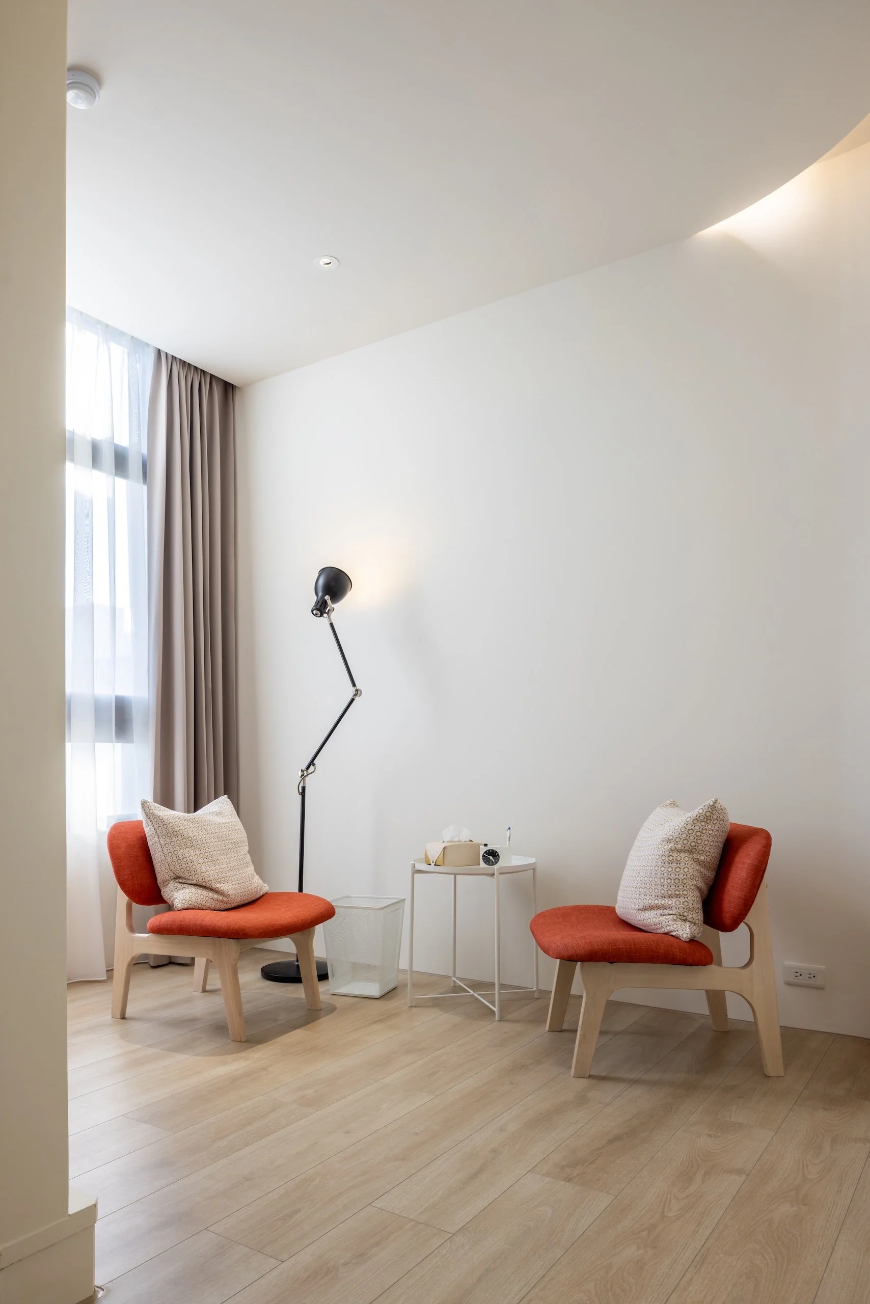 A cozy sitting area with two orange chairs, each with patterned cushions, a small white side table with a tissue box, a black floor lamp, and a large window with sheer white curtains and beige drapes. The room has light wooden flooring and plain white walls.