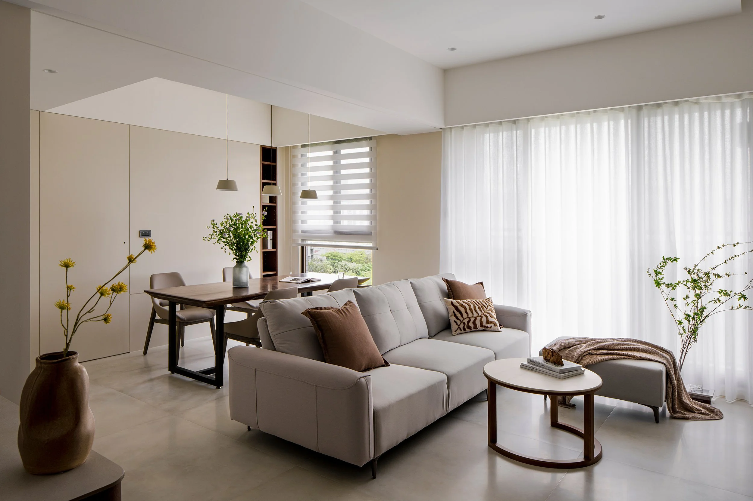 Modern living room with white sofa, brown and animal print throw pillows, small round coffee table with books, large windows with sheer curtains, dining area with wooden table, beige chairs, hanging pendant lights, potted plants, and a tall vase with yellow flowers.