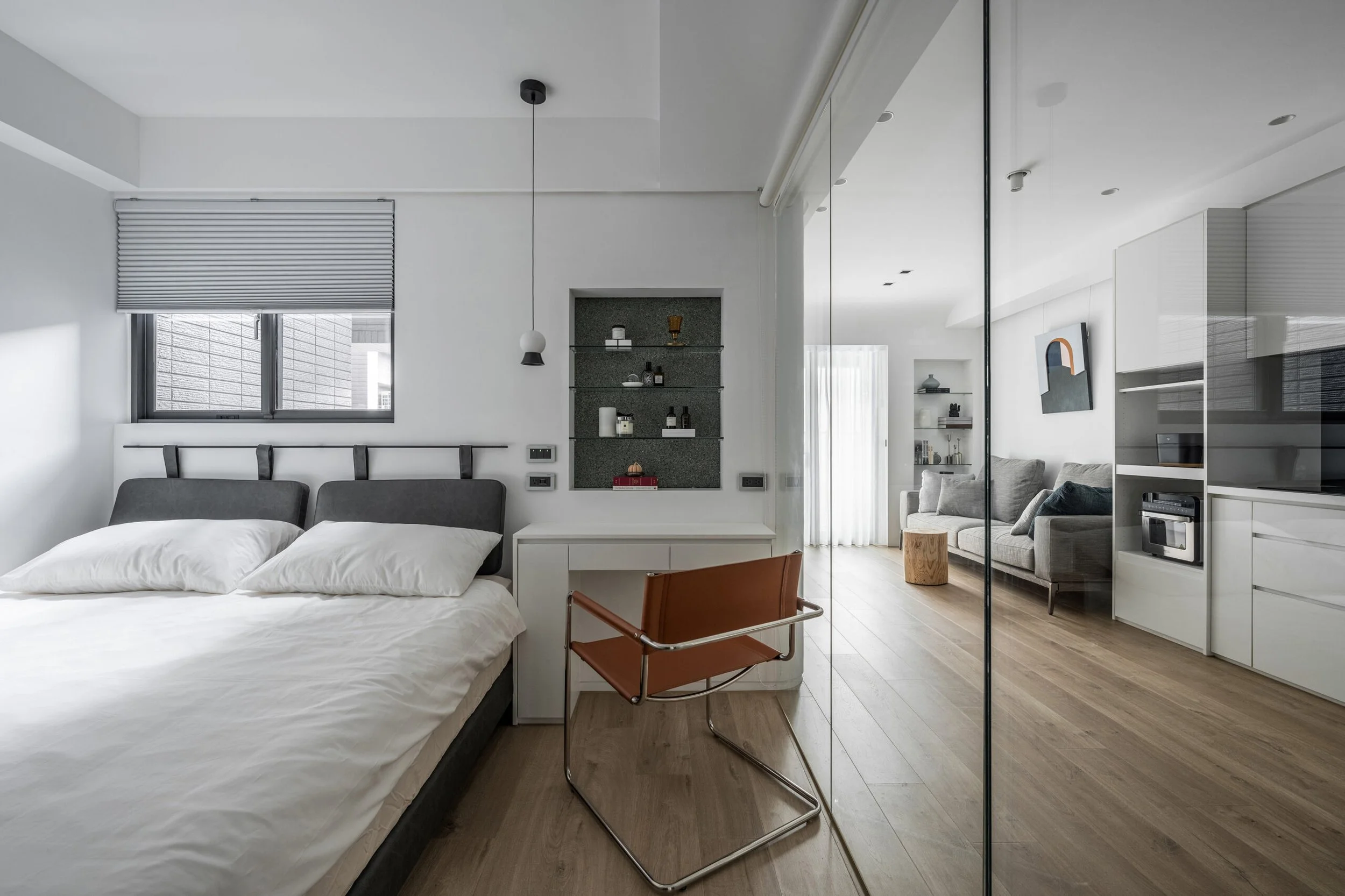 A modern, minimalist bedroom with a large bed, white bedding, and gray pillows. There is a window with a partially closed blind. An orange chair with a metal frame is next to a white desk. A gray built-in shelf with decorative items and bottles is above the bed's headboard. The room extends into a living area with a gray sofa, abstract art, and a mix of furniture and decor, separated by a glass wall.