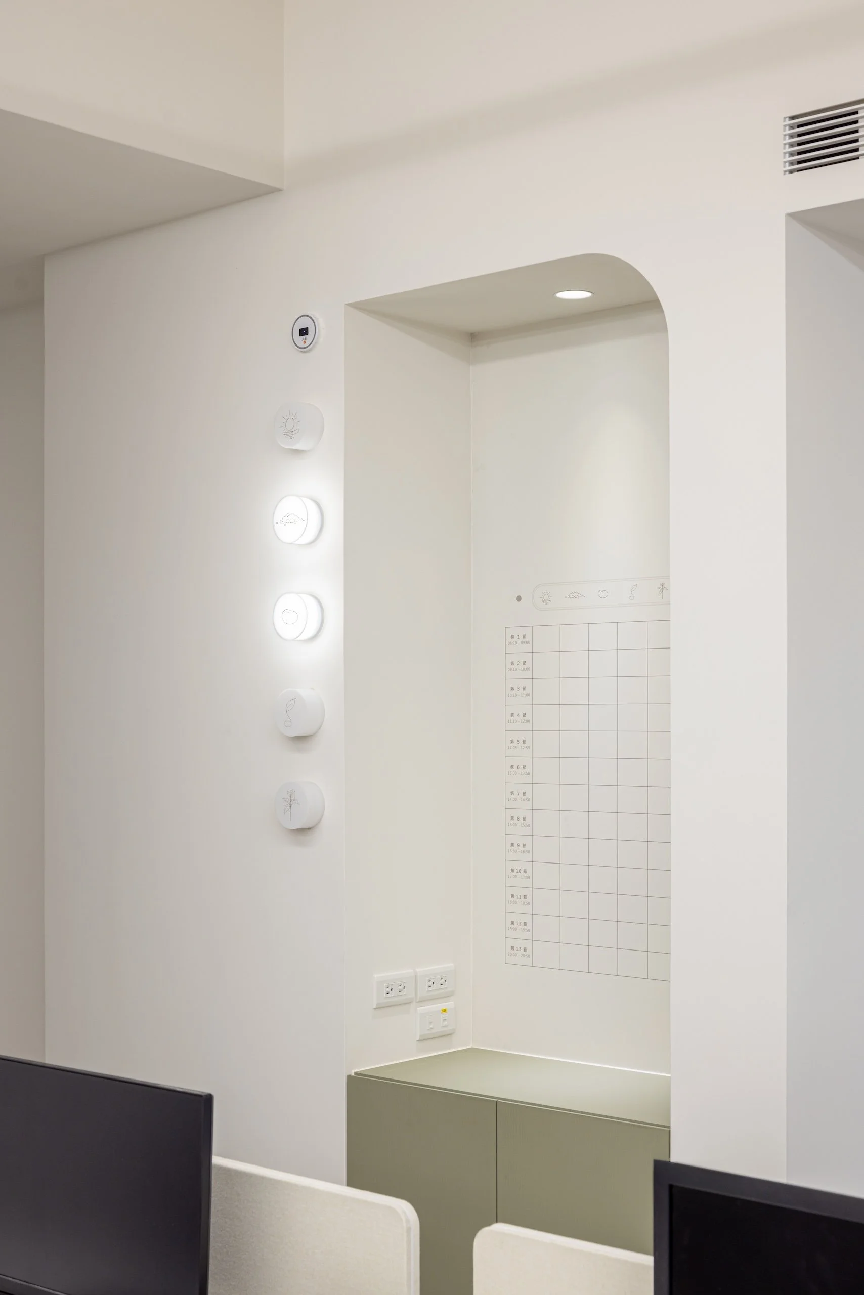 Interior of a modern office or reception area with white walls, a light-colored cabinet, and multiple computer monitors on desks. The wall features a whiteboard or schedule and wall-mounted controls or lighting switches.