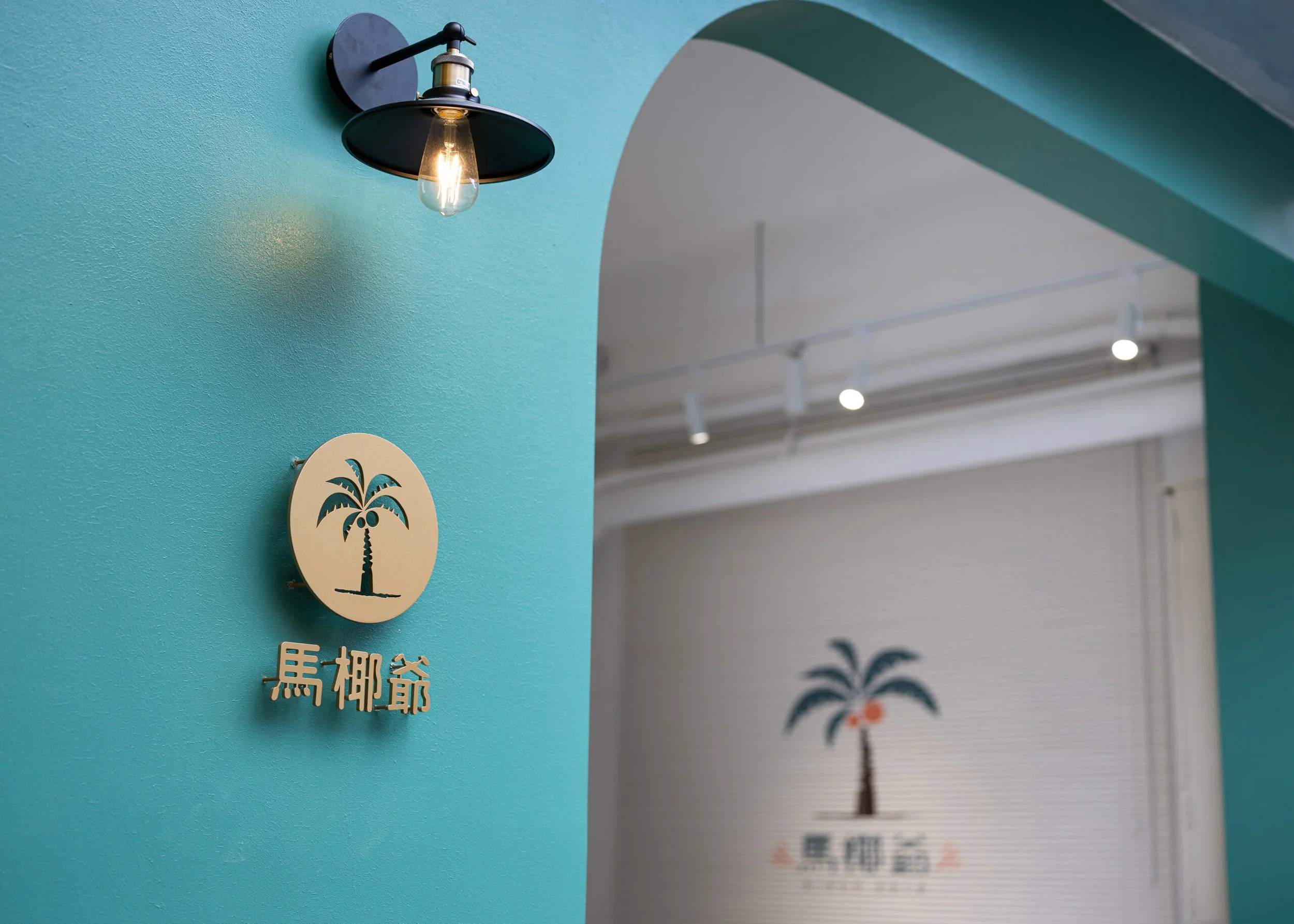 Close-up of an interior wall with a black wall-mounted light fixture, a decorative wooden plaque with a palm tree and Japanese text, and a partial view of a sign with a palm tree graphic and more Japanese text. The wall is teal, and the ceiling has exposed white pipes and track lighting.