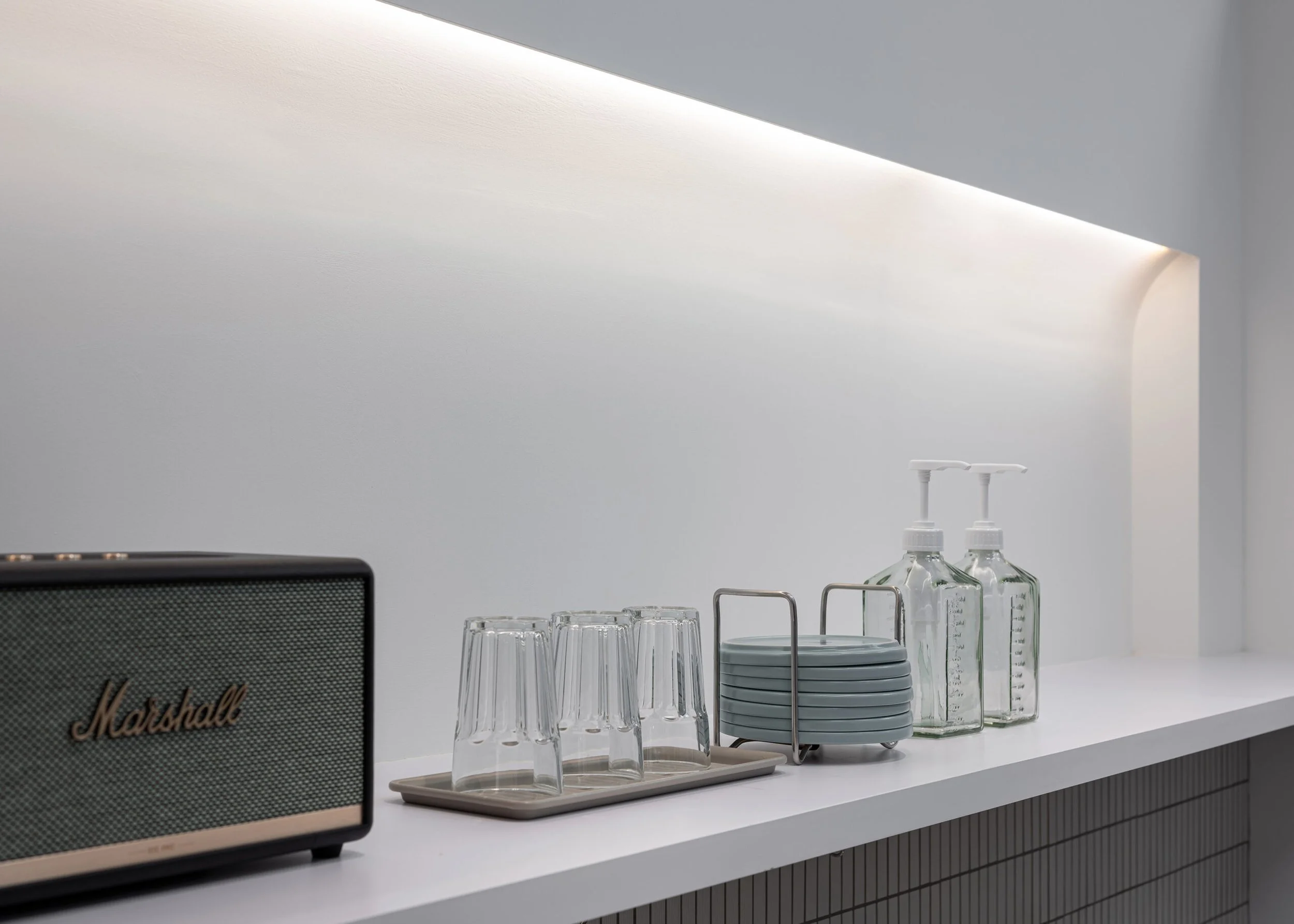 A minimalist kitchen or bar area with a white countertop, a stack of gray plates, clear drinking glasses, glass bottles with pumps, and a gray Marshall speaker. The background features a smooth white wall with soft lighting and a corner with a curved edge.