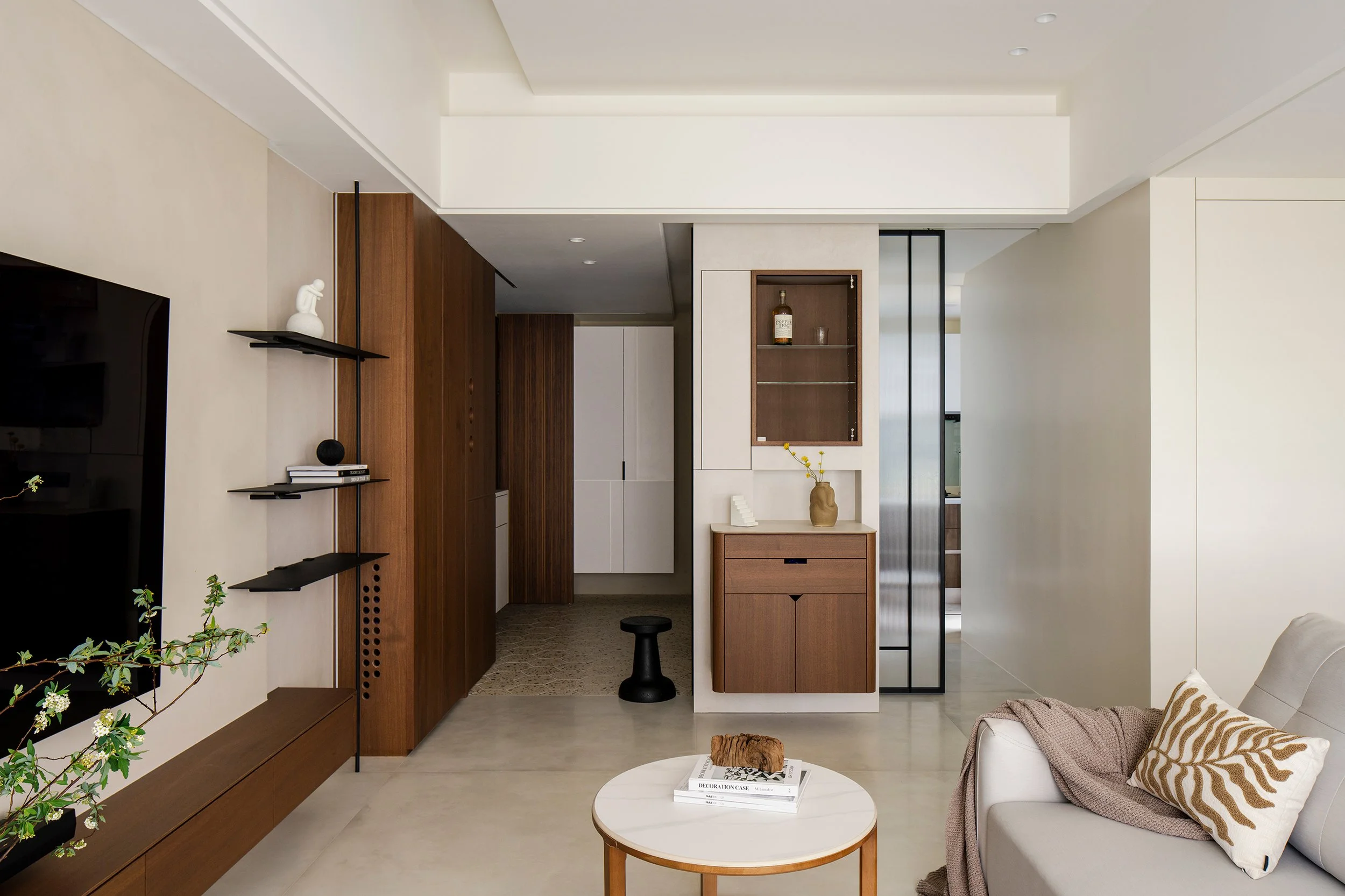 Modern living room with beige walls, a white ceiling, and wooden accents, featuring a beige sofa with patterned cushions, a round coffee table with books and a decorative item, a black stool, and built-in wooden and white cabinets.