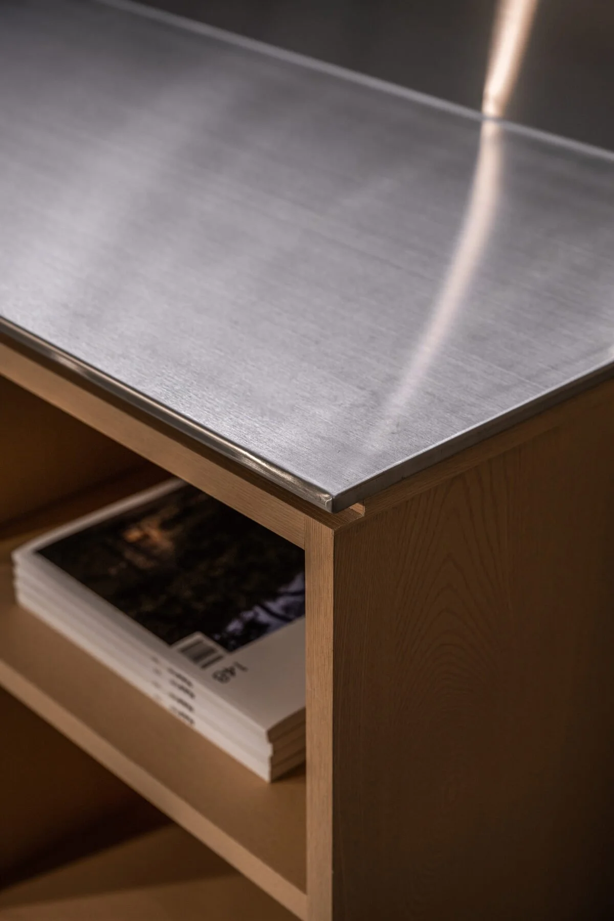 Close-up of a wooden side table with a metal top, a magazine or brochure is visible on the lower shelf.