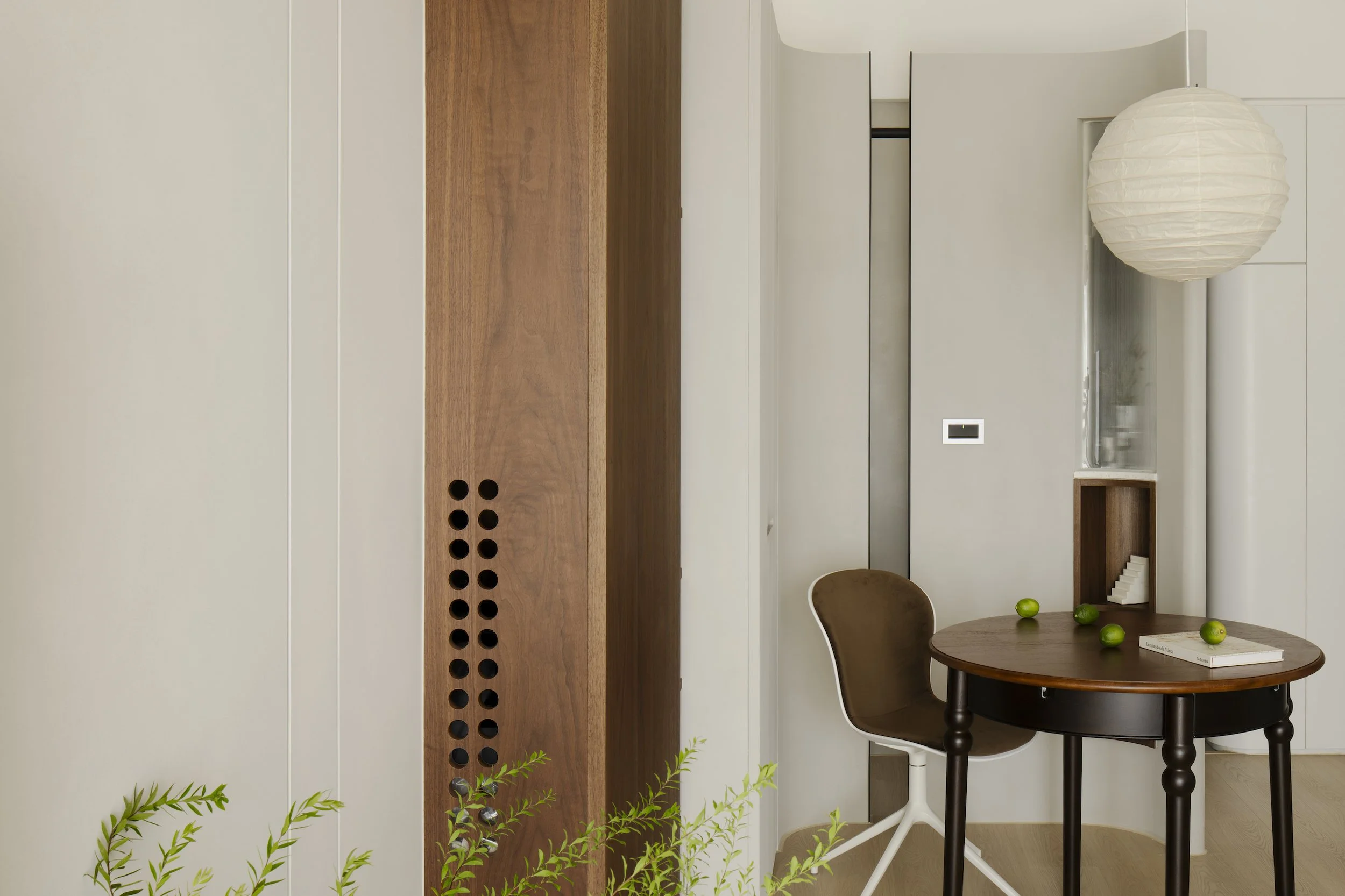 A modern interior with white wall panels, a wooden accent panel with black circular vents, a small round wooden table with green limes, a white and brown chair, and a white paper lantern pendant light.