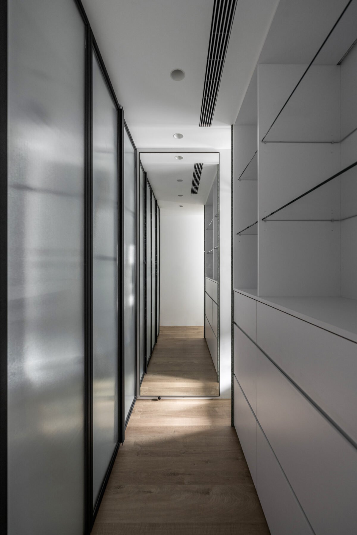 A narrow hallway with frosted glass closet doors on the left and white storage cabinets with open shelves on the right. A large mirror at the end reflects the hallway, with ceiling air vents and recessed lighting.