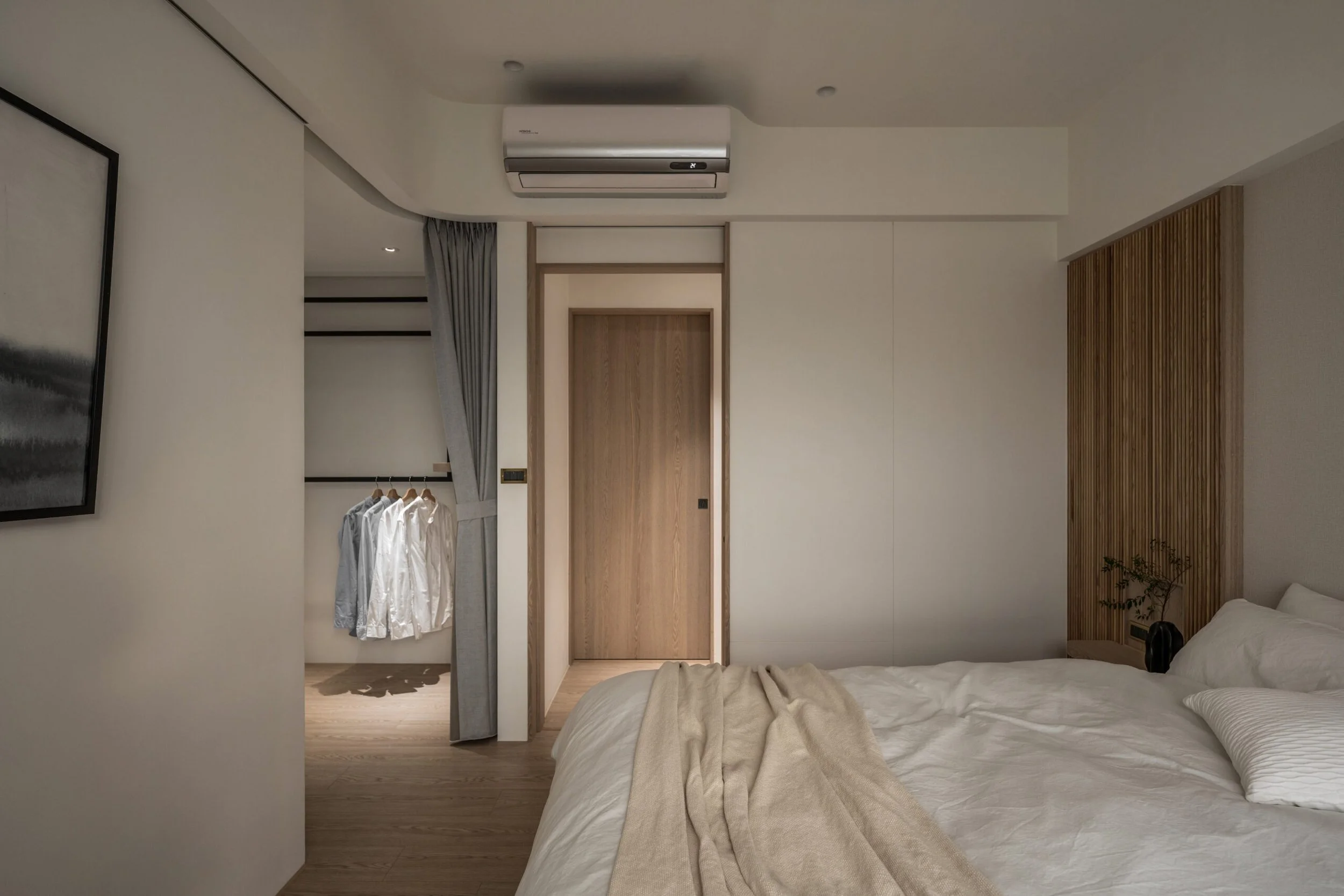 A modern bedroom featuring a bed with white bedding and pillows, a wooden sliding door, and a built-in closet with clothes hanging. The room includes a wall-mounted air conditioner, a small black item on the bed, and a potted plant near the bed. There is a framed artwork on the wall and wooden accents on the wall.