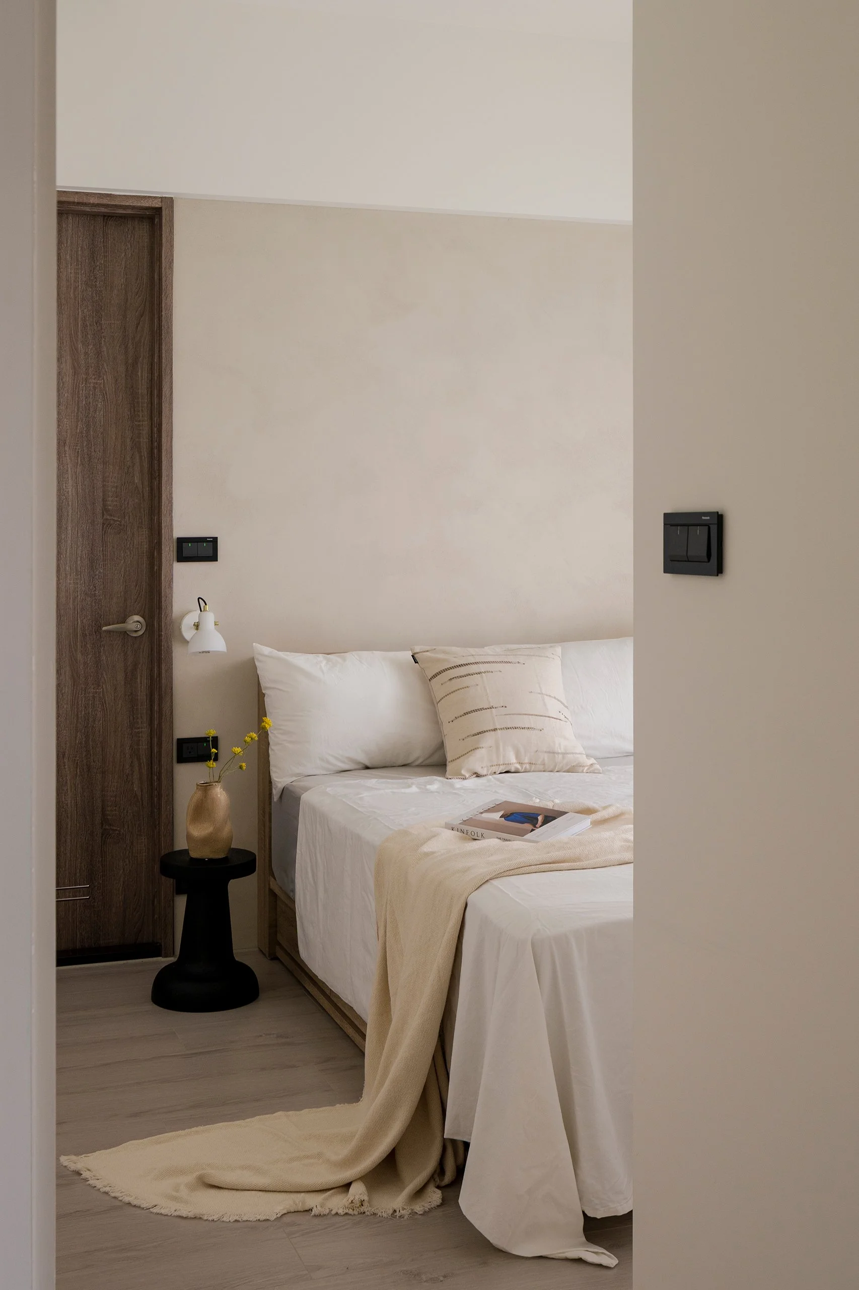 A cozy bedroom with two single beds, one with white bedding and a beige blanket, a black side table with a beige vase holding yellow flowers, a wooden door, a wall-mounted lamp, and light-colored wooden flooring.