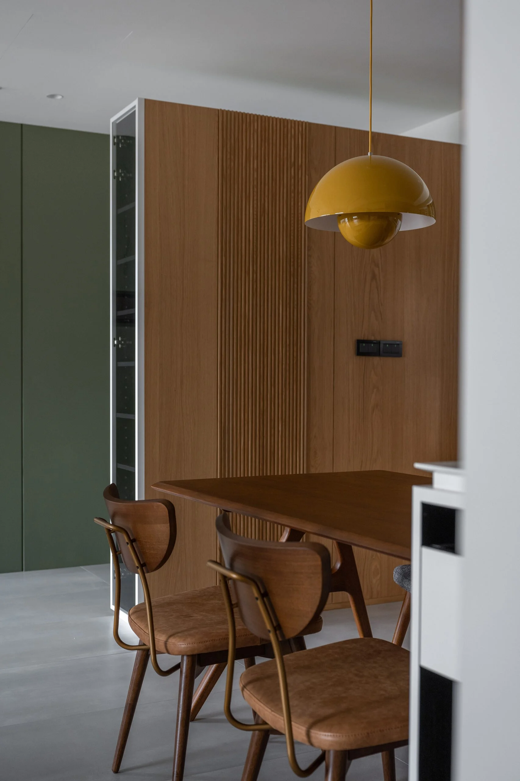 Interior view of a dining area with a wooden table and chairs, a wooden wall with vertical slats, a yellow hanging lamp, and a green cabinet.
