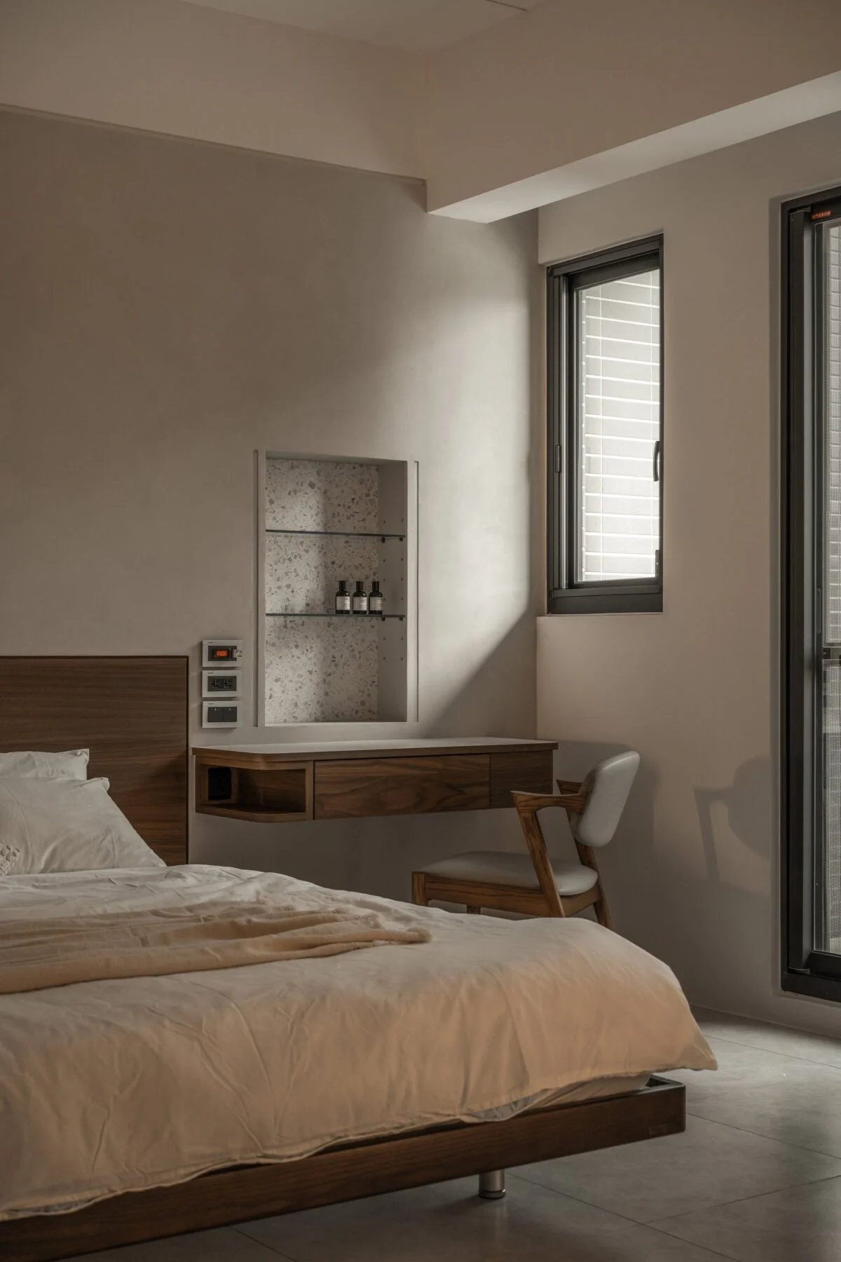 Modern bedroom with white walls, wooden bed frame with beige bedding, built-in shelf with bottles, and a wooden desk with a white chair near a window with horizontal blinds.