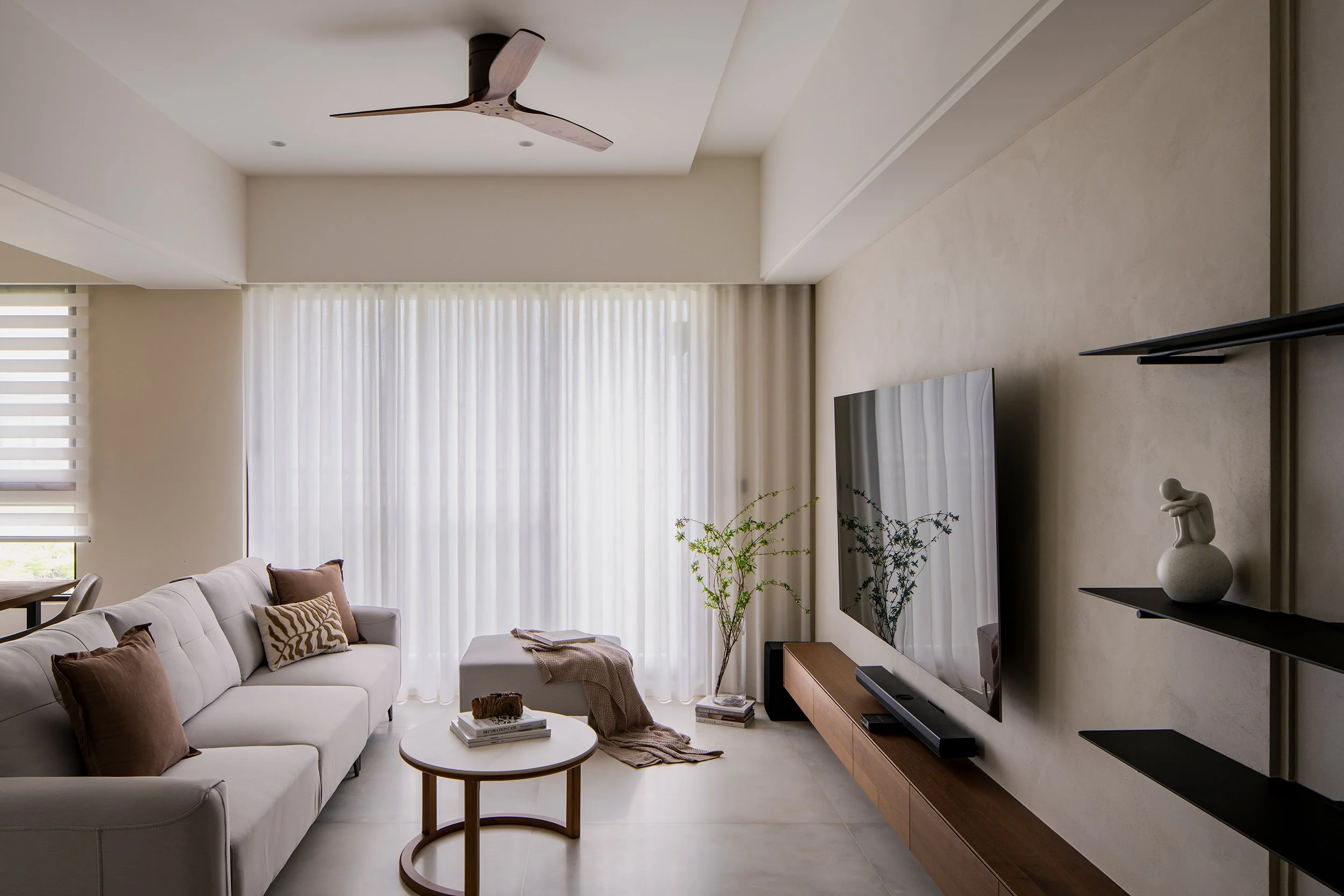 A modern, minimalist living room with a white sofa, brown pillows, a round wooden coffee table, a television on a sleek wooden and black shelving unit, and sheer white curtains letting in natural light.