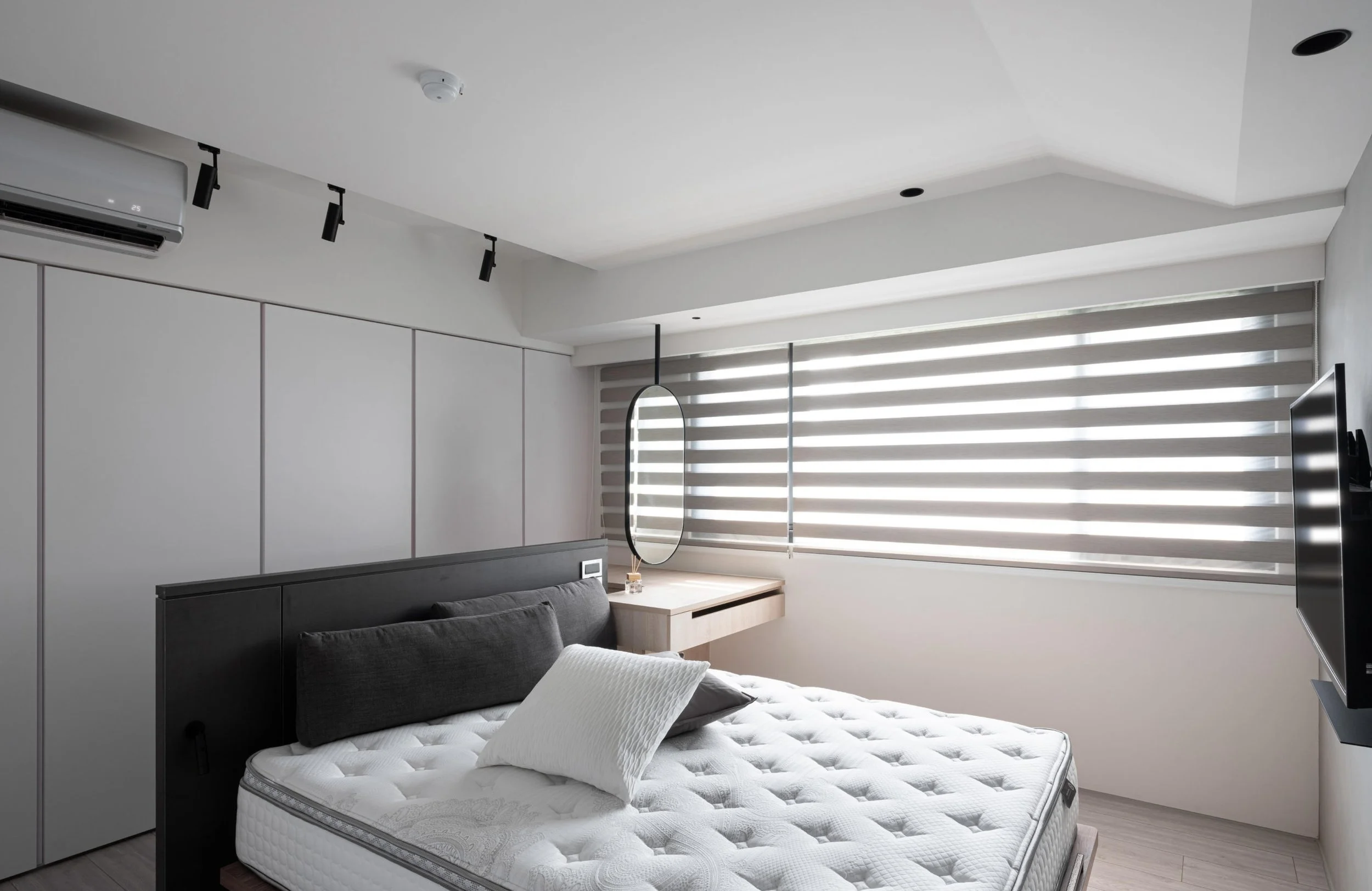 Modern bedroom with white walls, horizontal blinds on the window, and a bed with a mattress and pillows. A small desk with a mirror hangs above it, and a flat-screen TV is mounted on the wall.