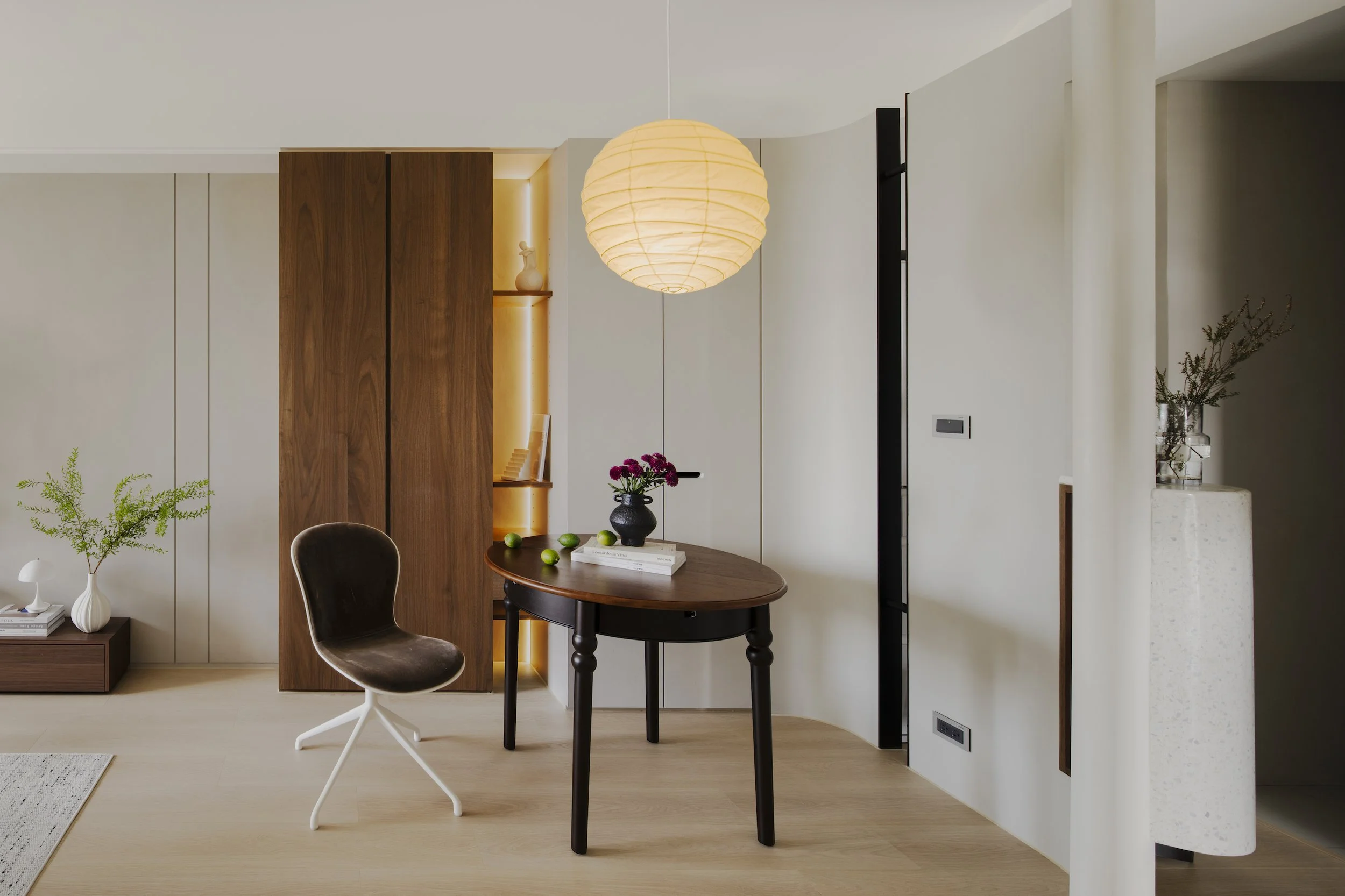 Modern dining room featuring a round wooden table with a black plant stand and a dark brown chair, a large paper pendant light, a white and wooden cabinet, greenery, and decorative items on shelves.