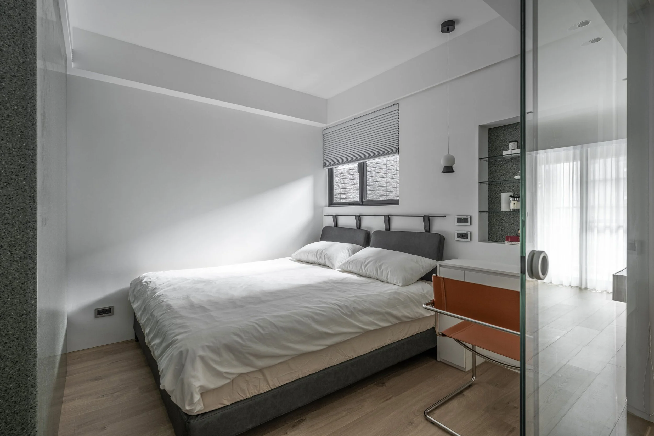 Modern bedroom with white walls, a window with blinds, a gray headboard, and a bed with white linens. There is a black ceiling pendant light, wall electrical outlets, and a glass wall or door with a metal handle. A small orange cabinet is beside the bed.
