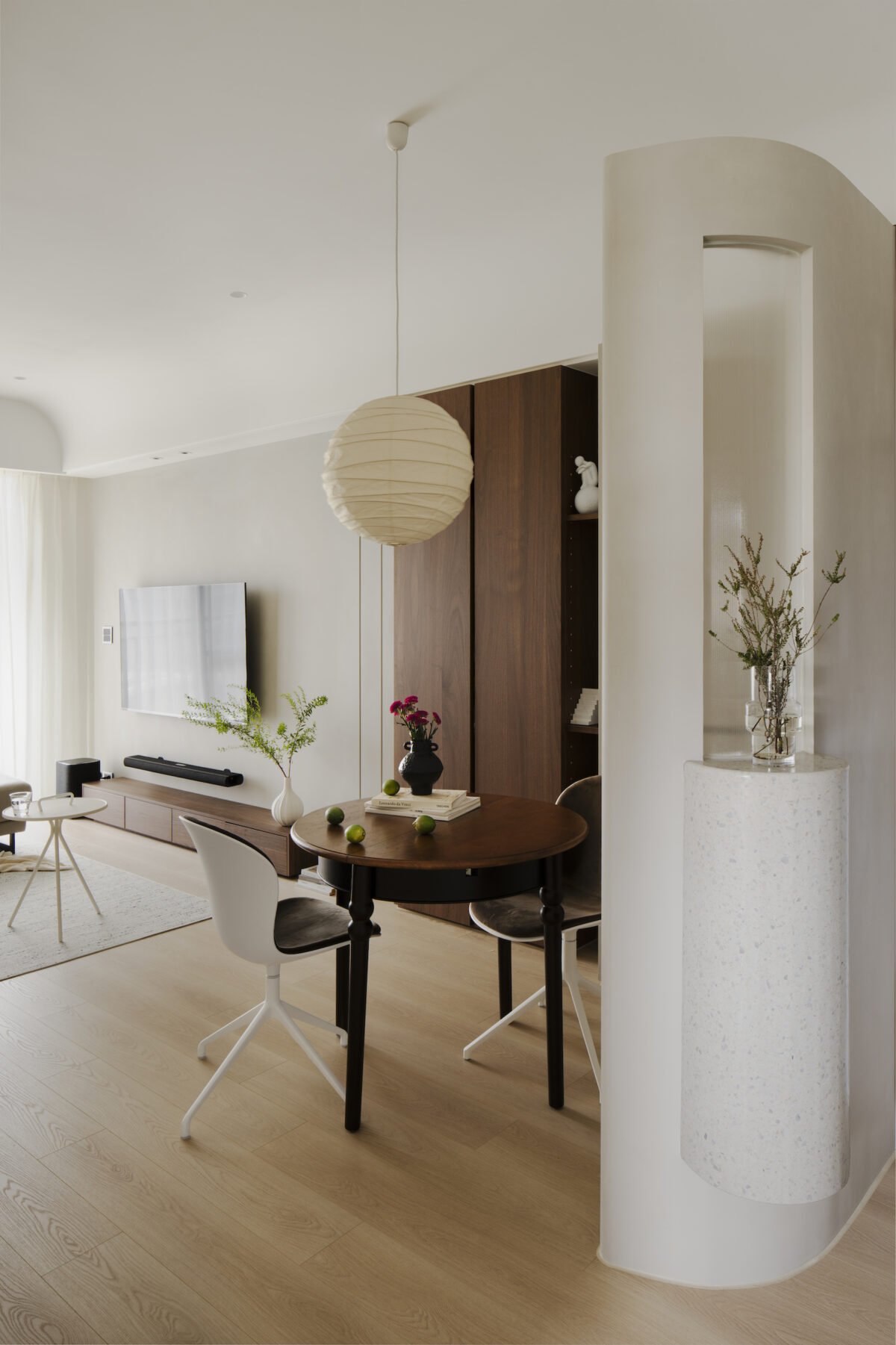 A modern living room with a round wooden dining table, white chairs, a beige rug, a wall-mounted TV, a wooden cabinet, and decorative plants.