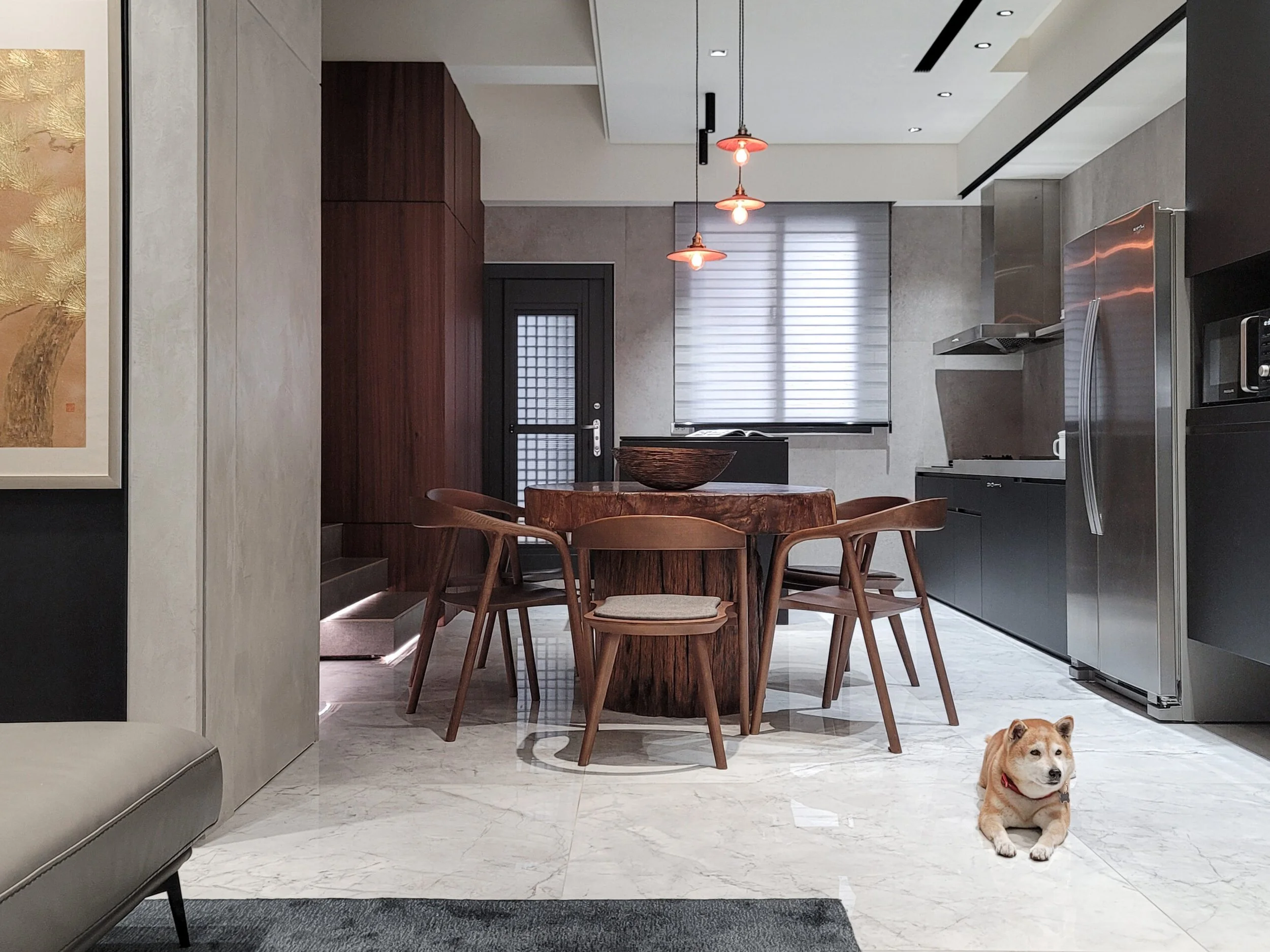 Modern kitchen and dining area with marble flooring, wooden dining table with matching chairs, pendant lights hanging from the ceiling, black door with glass panel, stainless steel refrigerator, microwave, and oven. A dog is lying on the floor in the foreground.