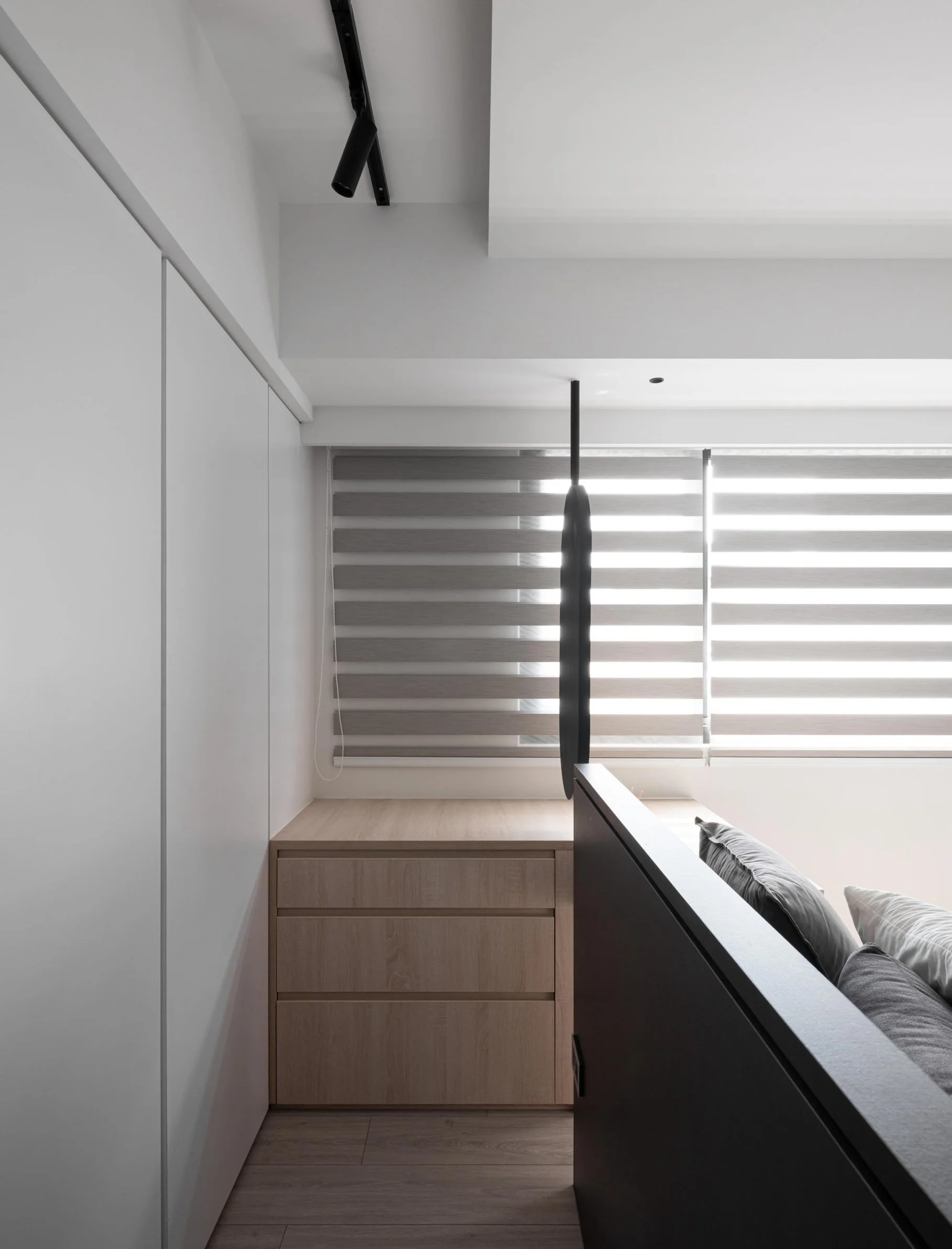 A minimalist bedroom with a black pendulum-style lighting fixture, wooden dresser, floor-to-ceiling window with horizontal blinds, and a bed with gray and white pillows.