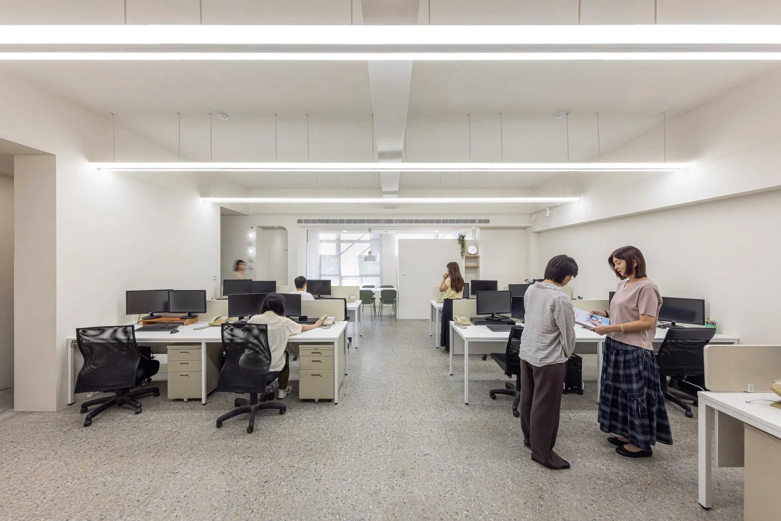 An open office space with workers using computers, two women standing and chatting, and natural light coming through large windows.