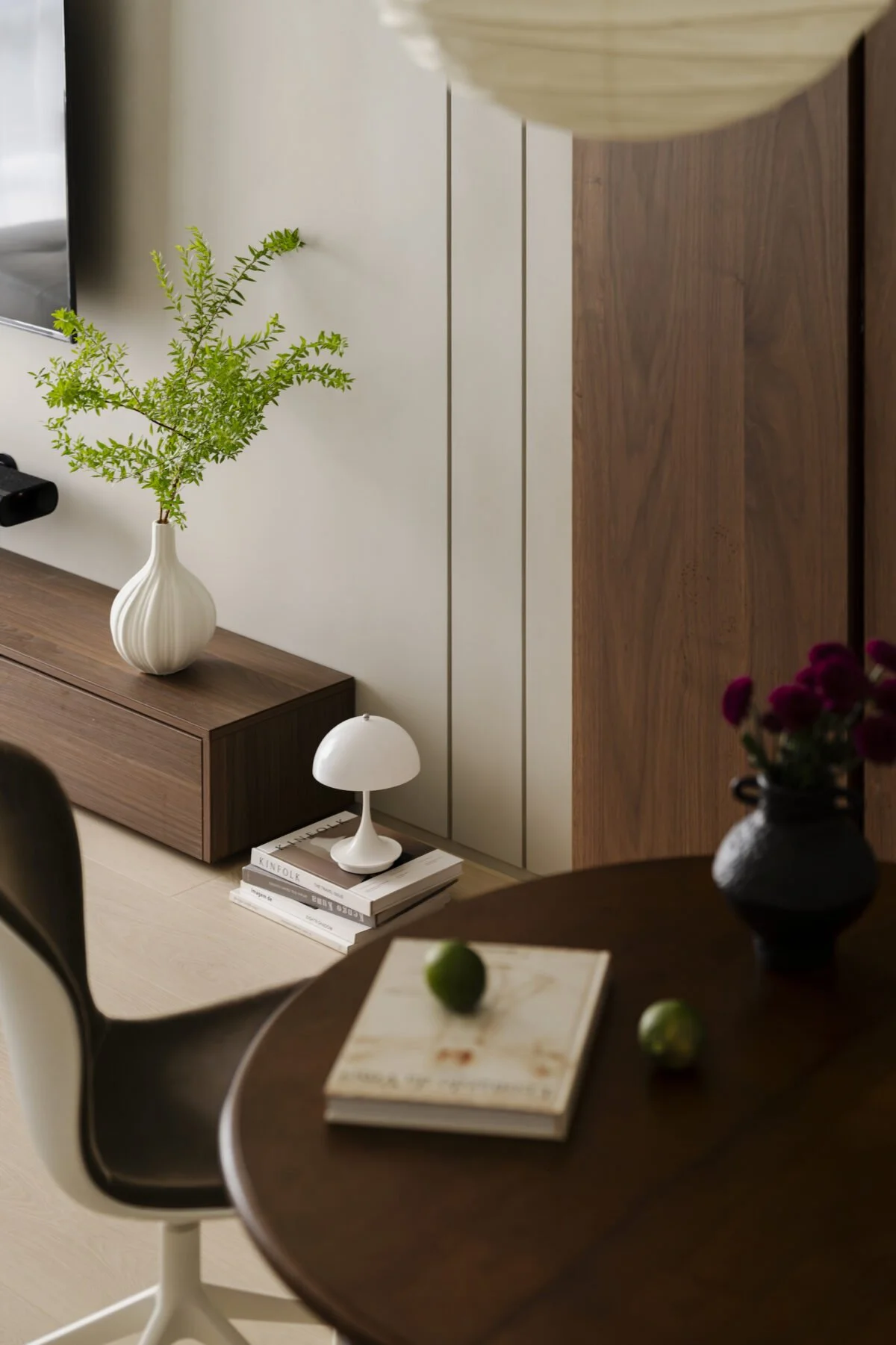 Interior of a room with a wooden cabinet, a white vase with green foliage, a small white table lamp on a stack of books, a wooden table with a dark finish, and some green fruits on a book in the foreground.