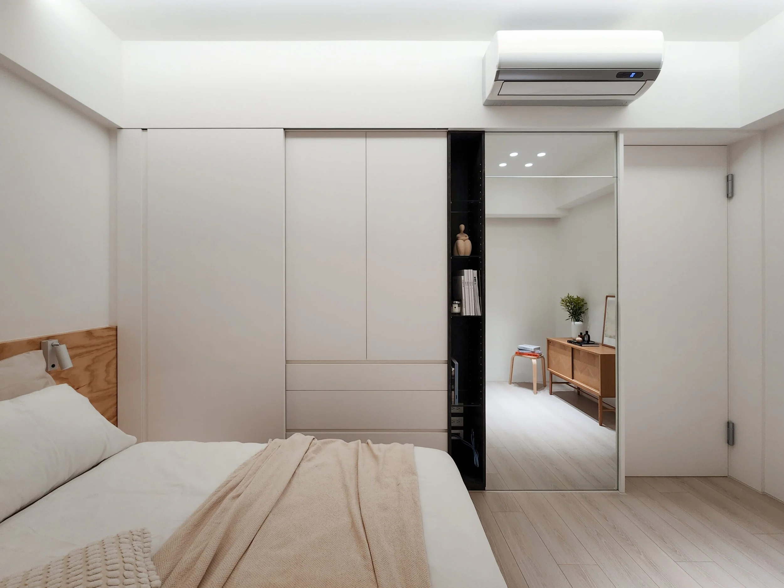 A bedroom with a white bed in the foreground, a black shelf with decorative items and books, a white wardrobe, and an adjacent room visible through an open doorway featuring modern furniture and decor.