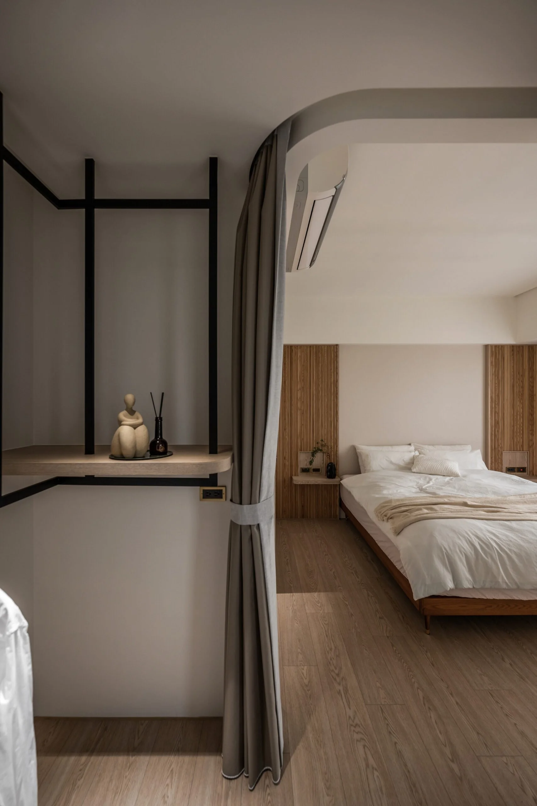 A bedroom with a double bed, beige bedding, and wood-paneled walls, framed by a gray curtain and partial view of a black metal shelving unit with decorative objects.