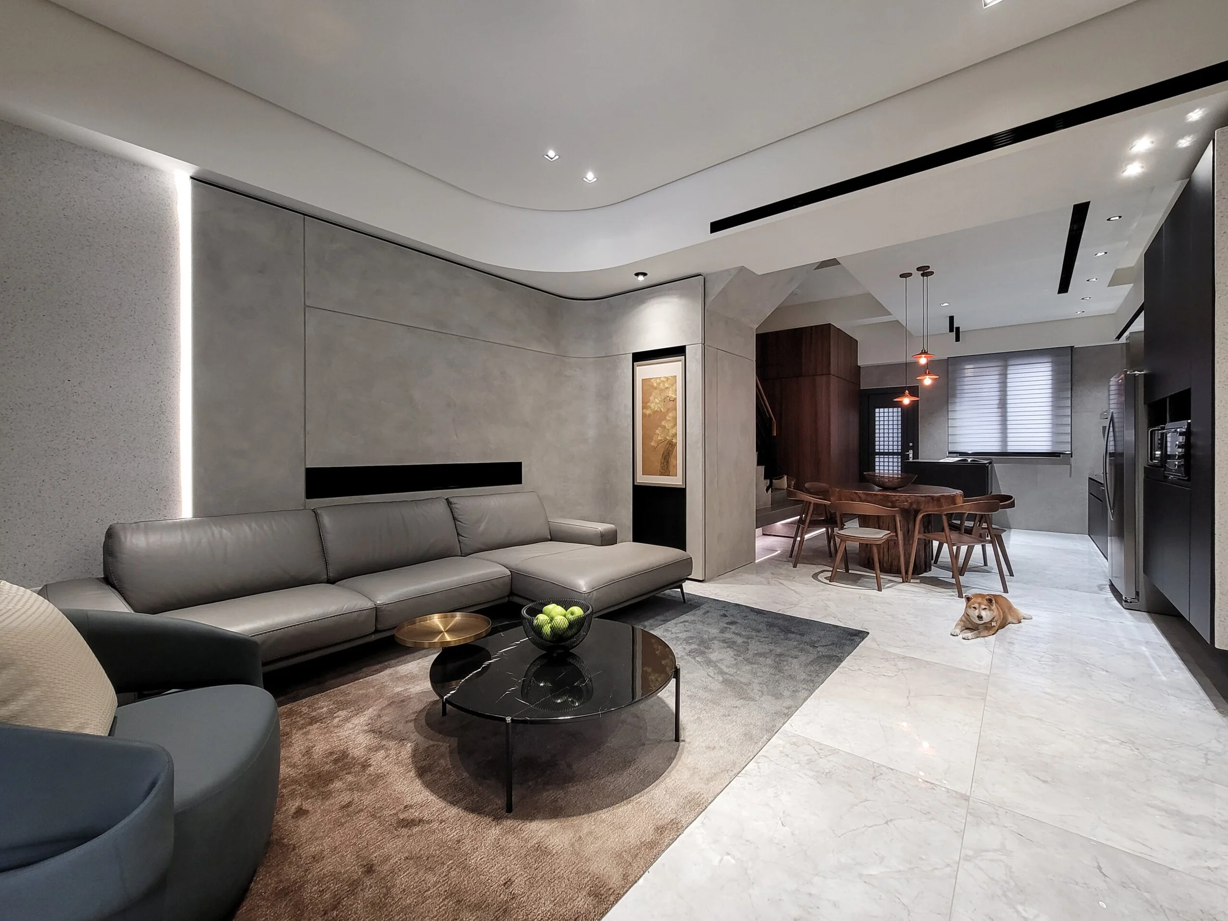 Modern living room with gray sofa, black coffee table with green apples, and a dog lying on the marble floor near the kitchen area.