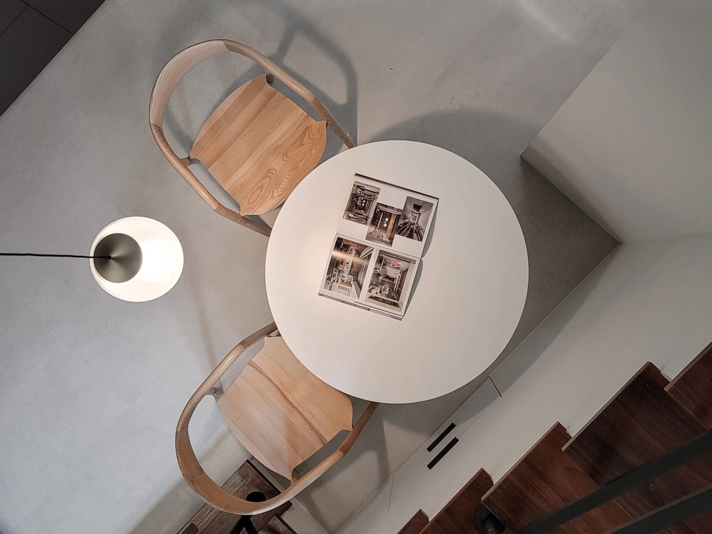 A white round table with a photo book open on it, two wooden chairs with curved backs, a modern white lamp with a black cord, on a gray floor with parts of a white wall visible.