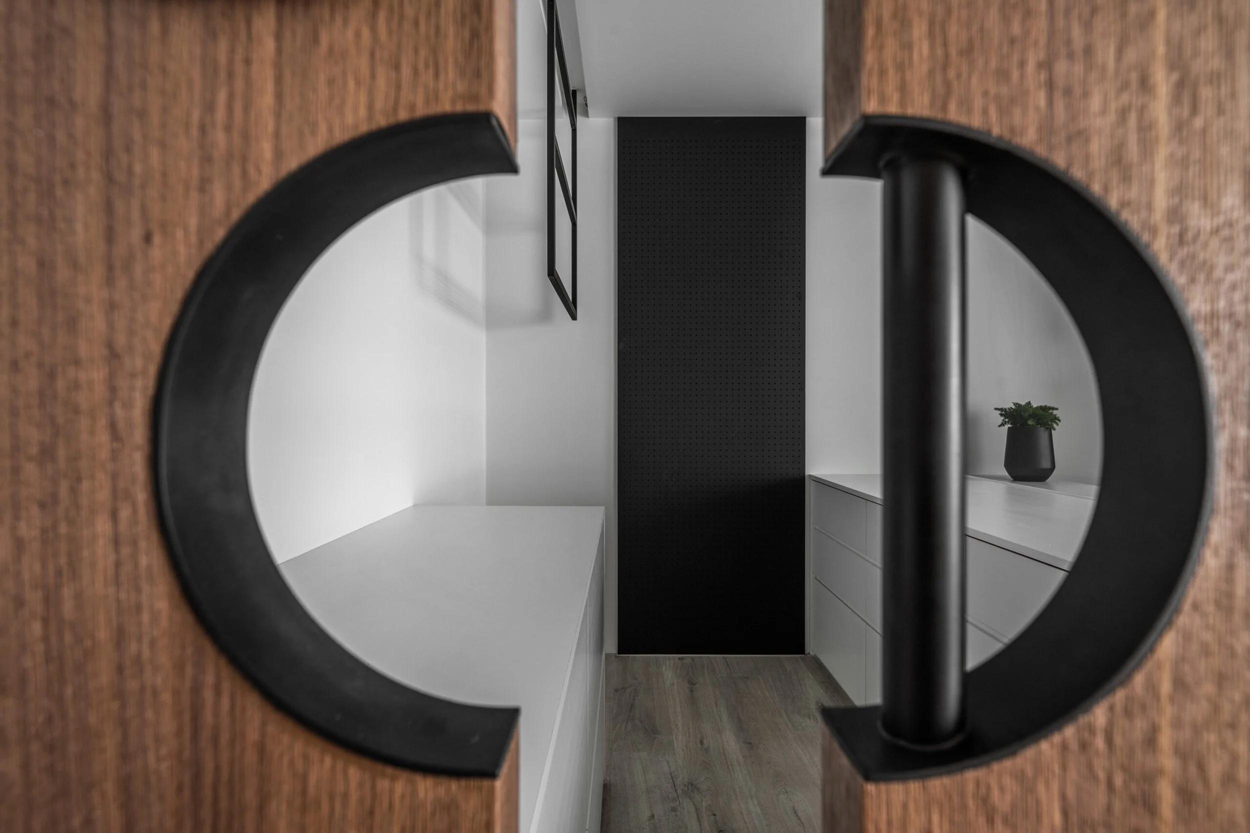 Interior view of a modern room seen through a circular mirror with a black frame. The room has white walls, a black pegboard on the back wall, a black framed shelving unit, a white cabinet with drawers, and a small black pot with a green plant.