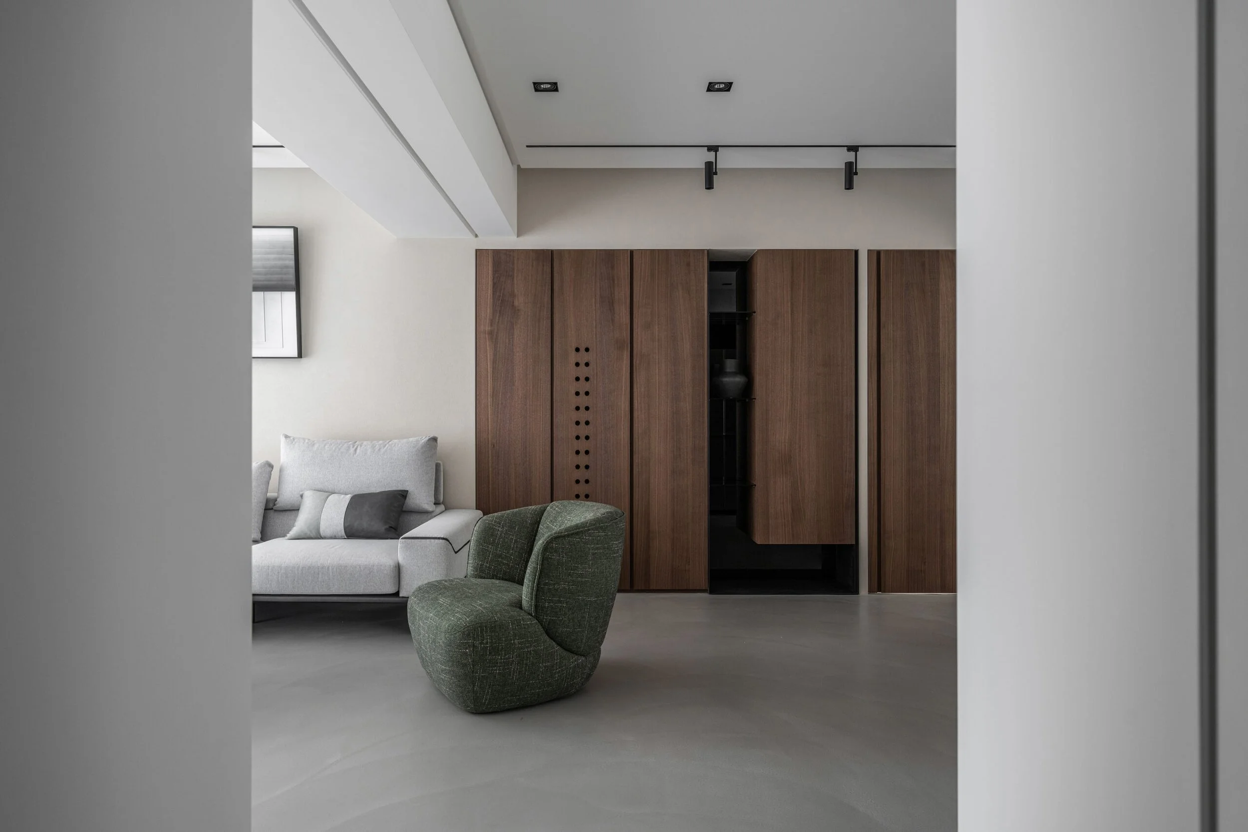 Minimalist living room with a white sofa and gray pillows, a green textured armchair, a wooden closet, a window with a roller blind, and black ceiling lights.