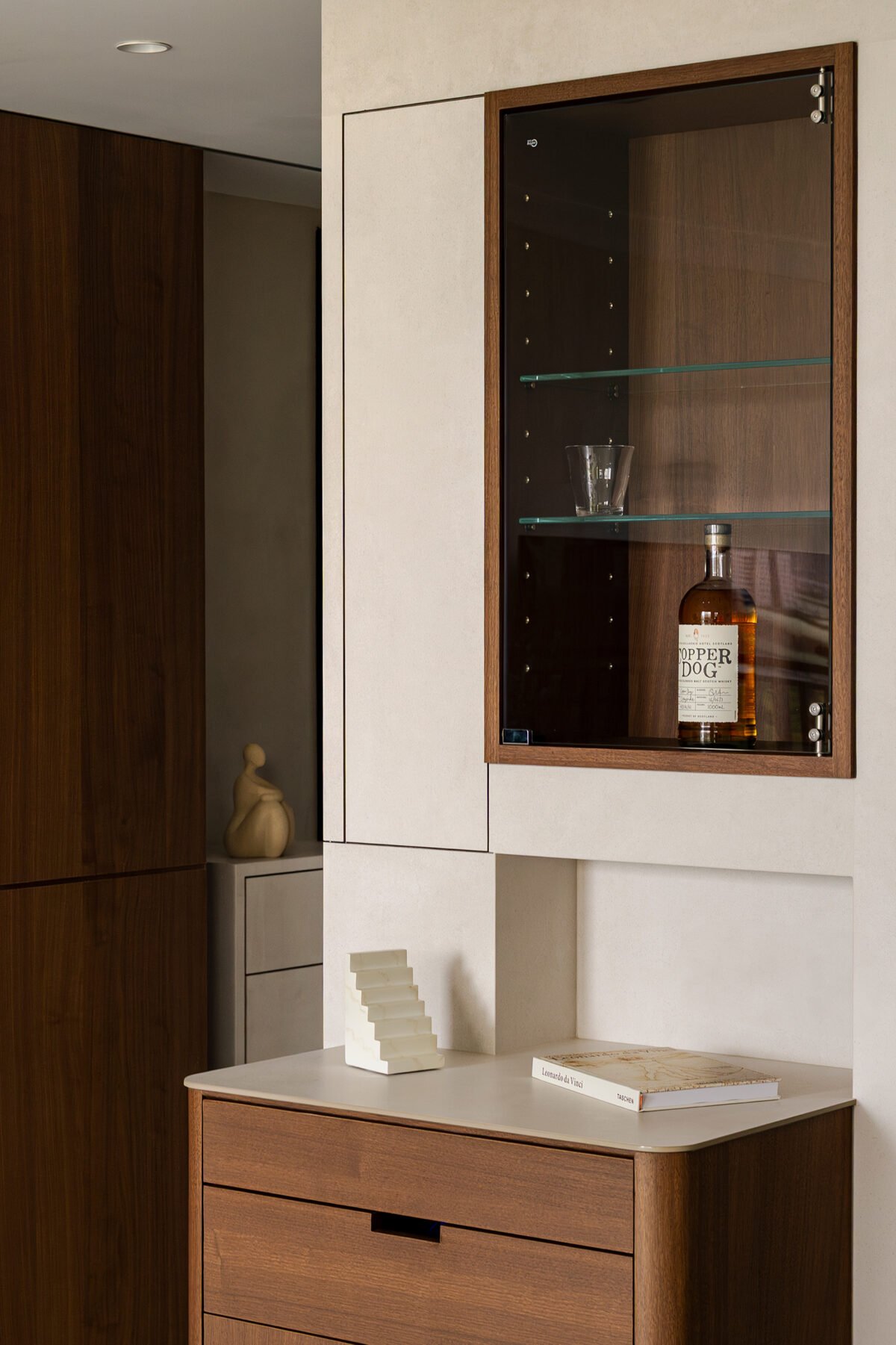 A wooden display cabinet with a glass door contains a bottle of liquor labeled "Copper Dog" and two glasses. The cabinet is mounted on a white wall, which also has a small white shelf below it with an abstract sculpture and a book. The room features a modern interior design with wood accents.
