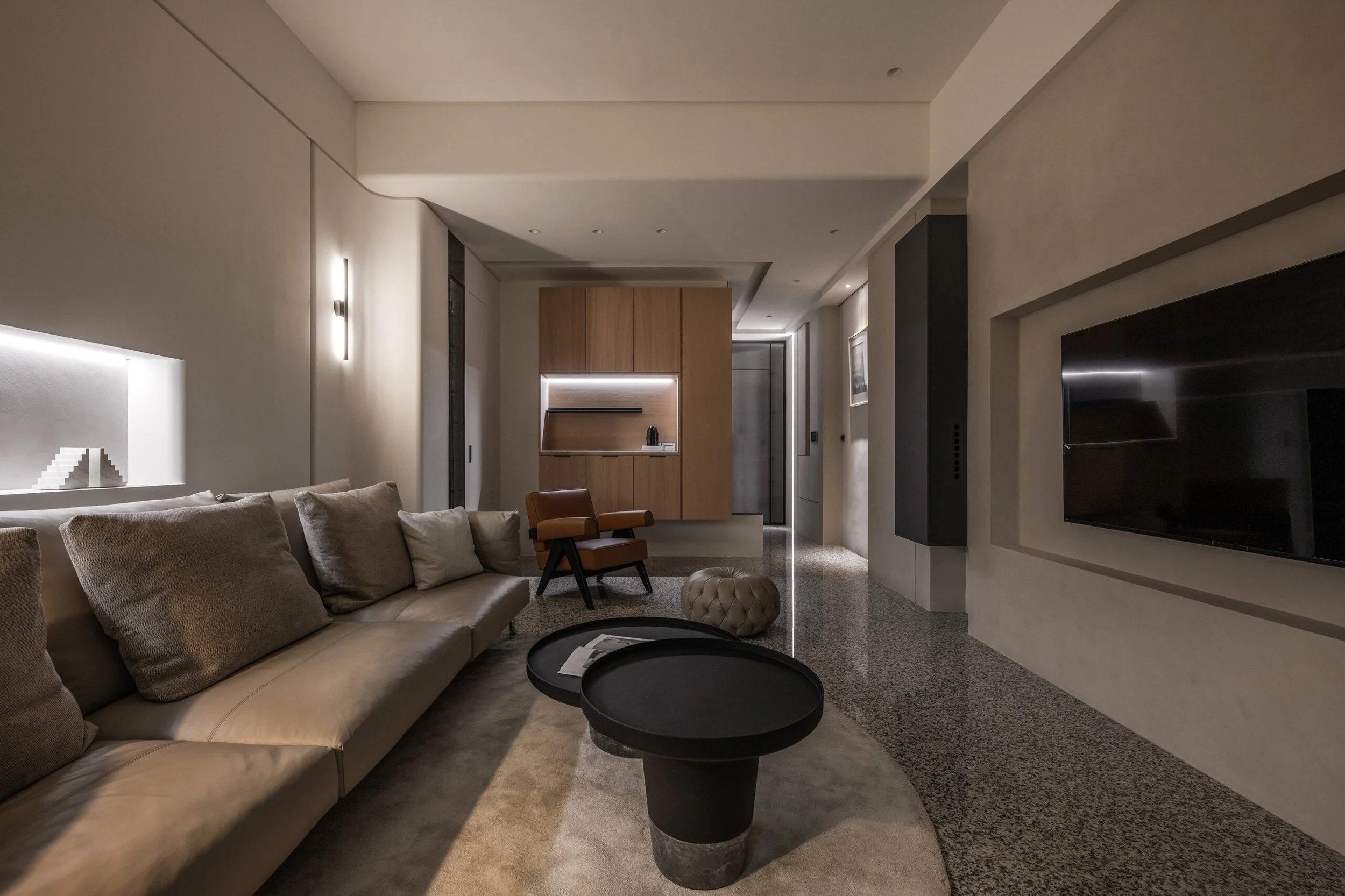 Modern living room with beige sofa, black round coffee tables, wall-mounted TV, wooden cabinet, and subtle lighting.