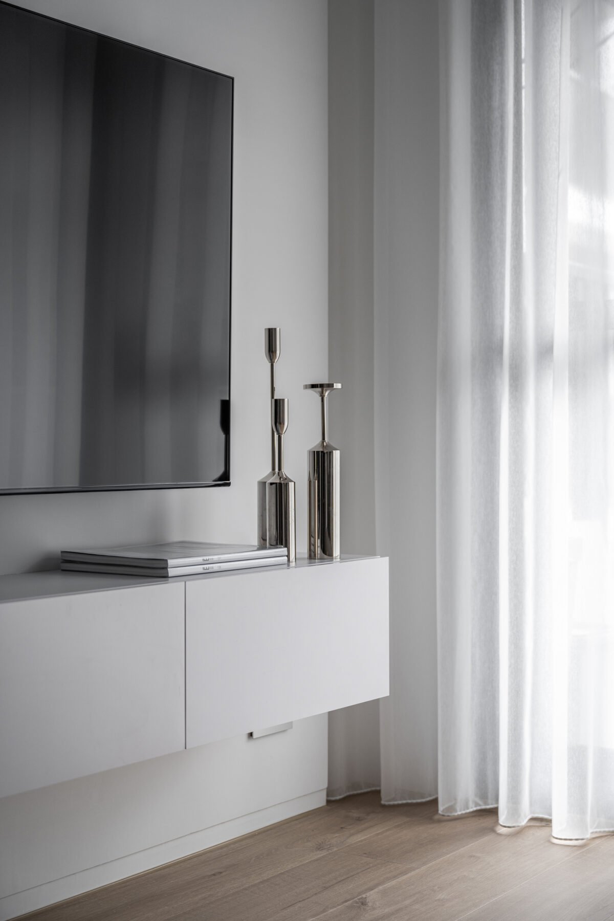 A minimalist living room corner with a large flat screen TV mounted on a white wall, a white floating cabinet below, and silver candle holders on top. Light curtain panels hang next to a window, allowing natural light to illuminate the space.