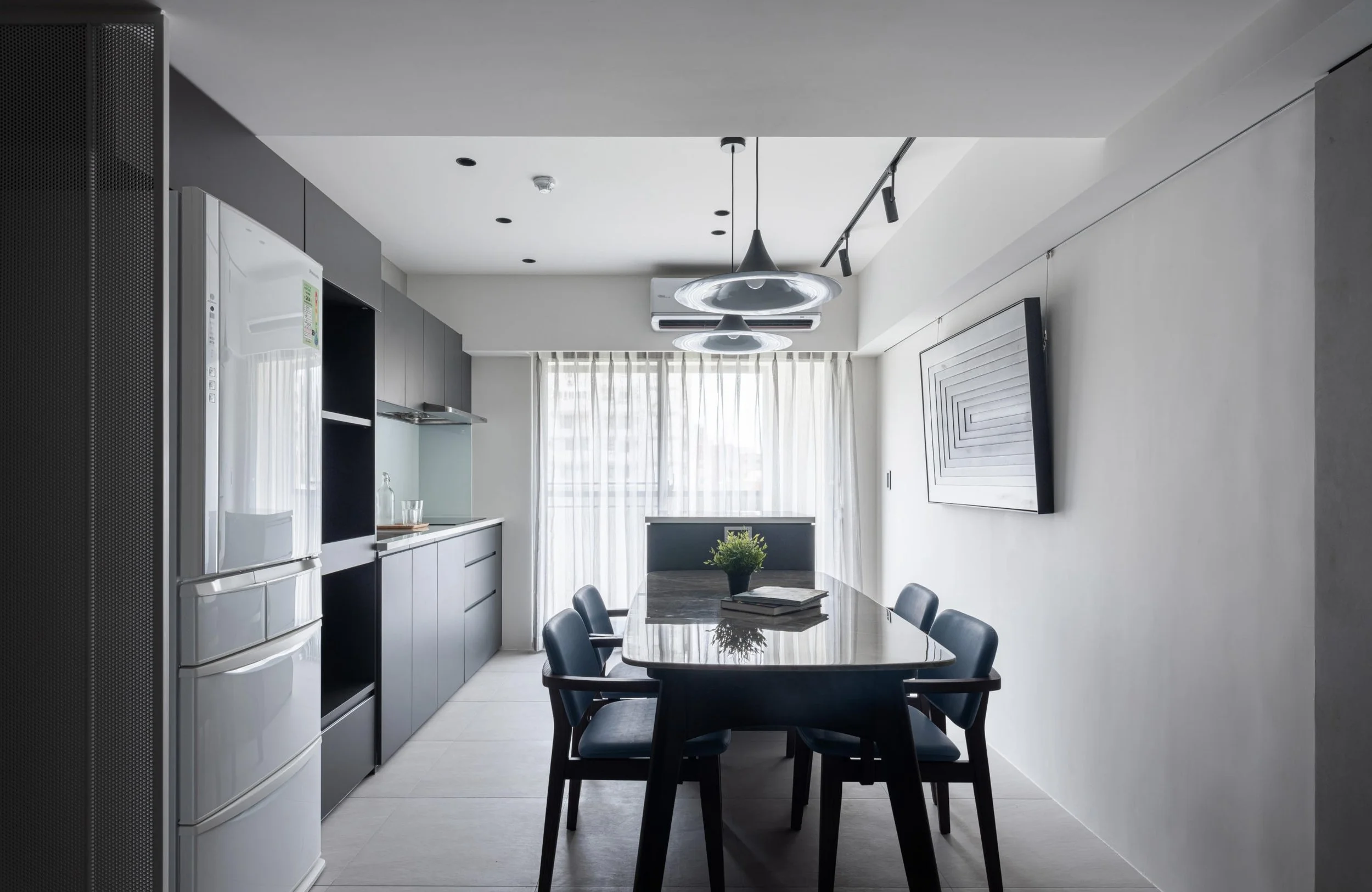 Modern dining room with a black dining table, eight black chairs, a gray kitchen with cabinets, a white refrigerator, a window with sheer curtains, ceiling track lighting, and a large framed artwork on the wall.