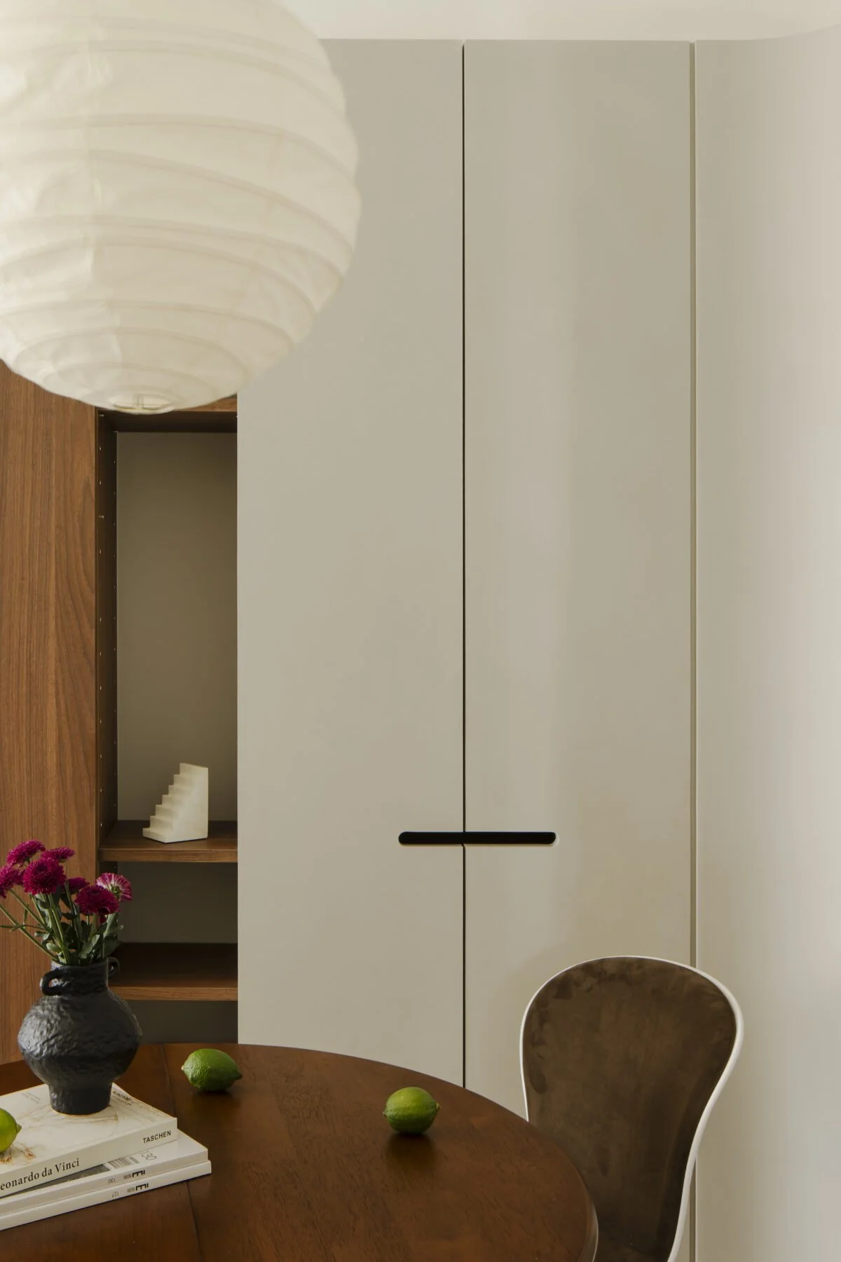Interior view of a dining room showing a wooden table with three limes, a black textured vase with pink flowers, and a couple of books. Part of a white, minimalist cabinet and a wooden shelf with decorative objects are visible in the background. A paper lantern hangs from the ceiling.