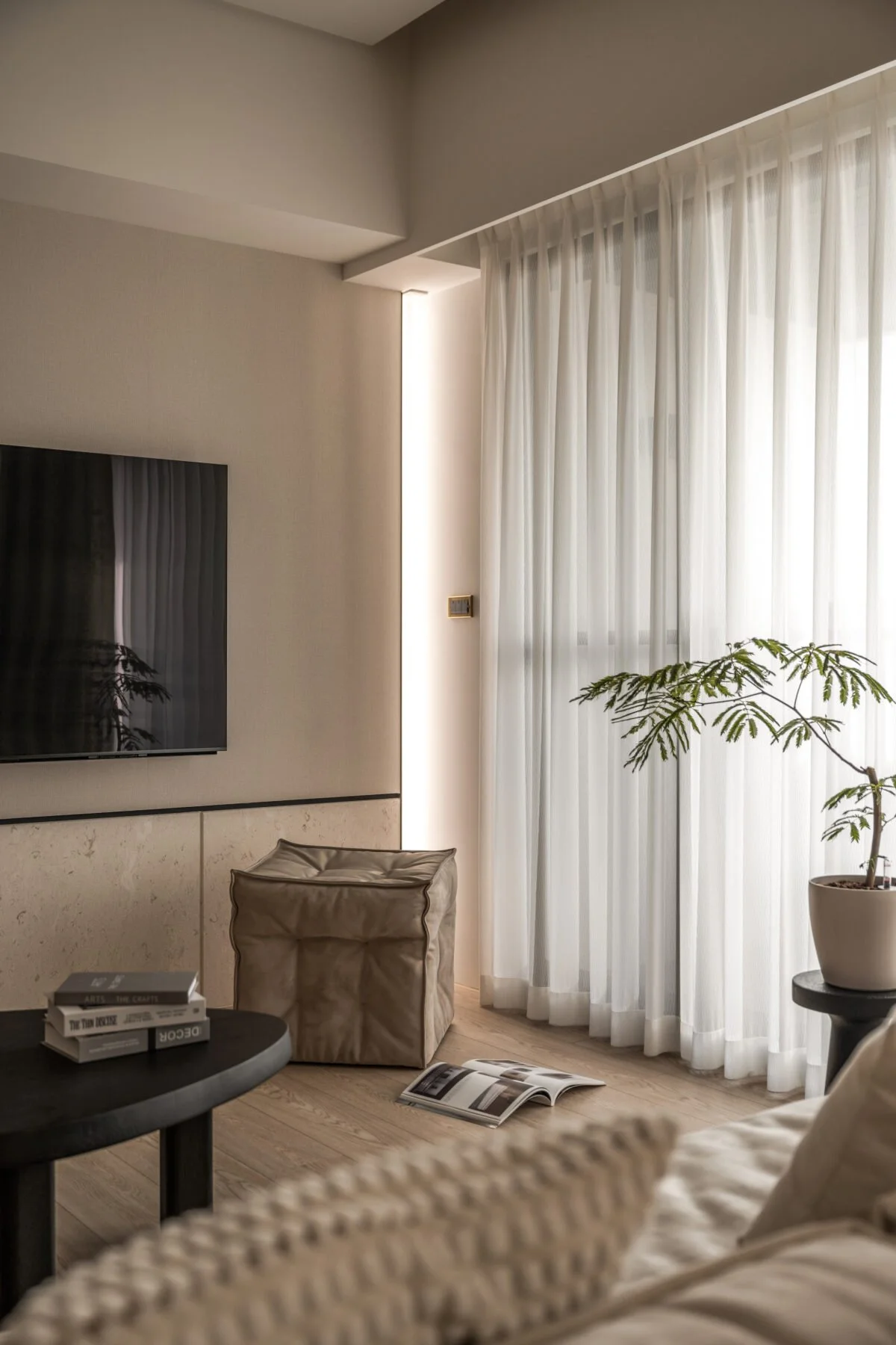 Living room with beige walls, white curtains, a flat-screen TV mounted on the wall, a potted plant, a round black table with books, and an upholstered armchair. An open magazine lies on the wooden floor.