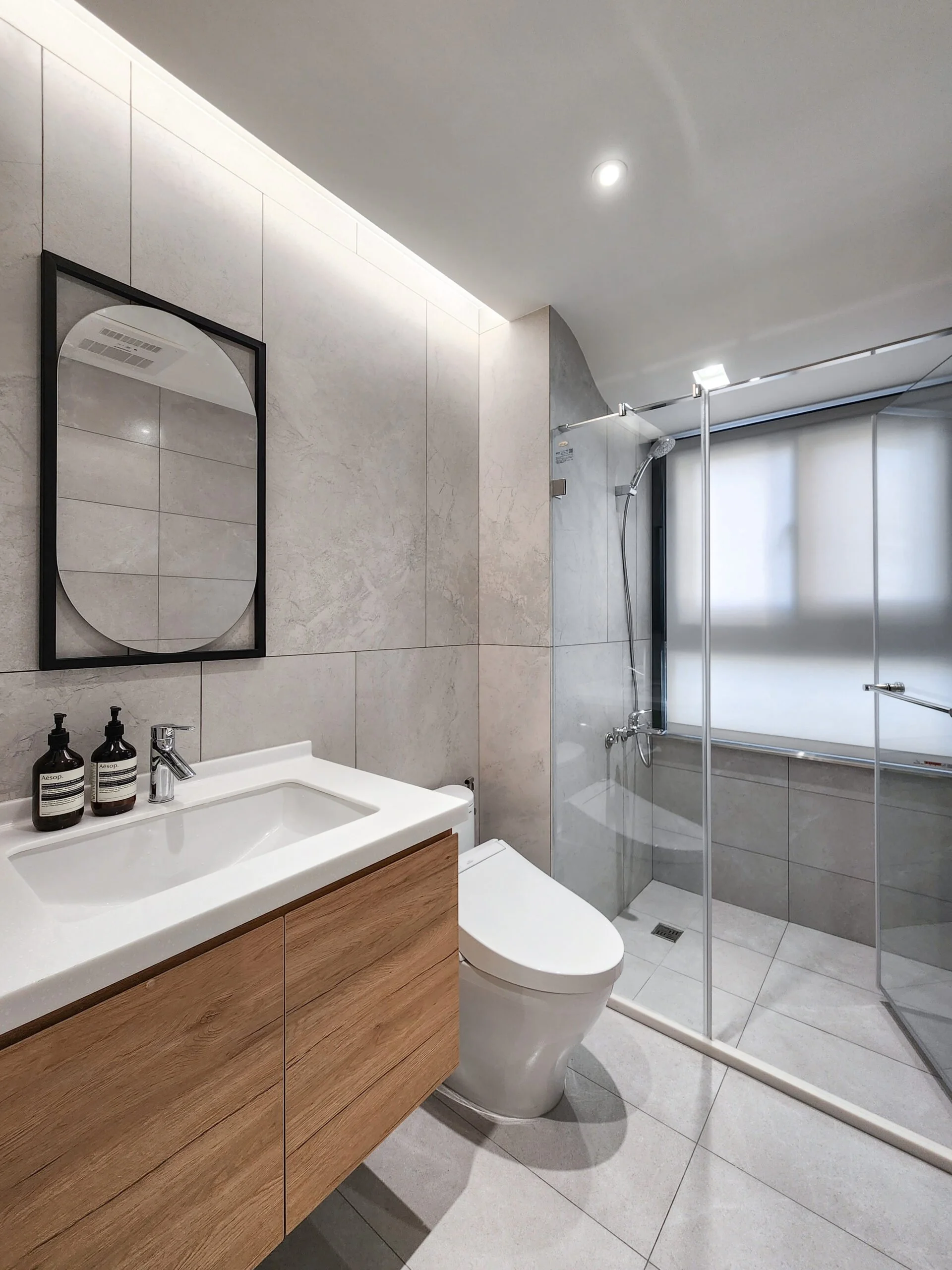 Modern bathroom with a wooden vanity, white sink, black framed mirror, toilet, and a walk-in shower with glass enclosure, tiled walls, and a showerhead, illuminated by ceiling lights.