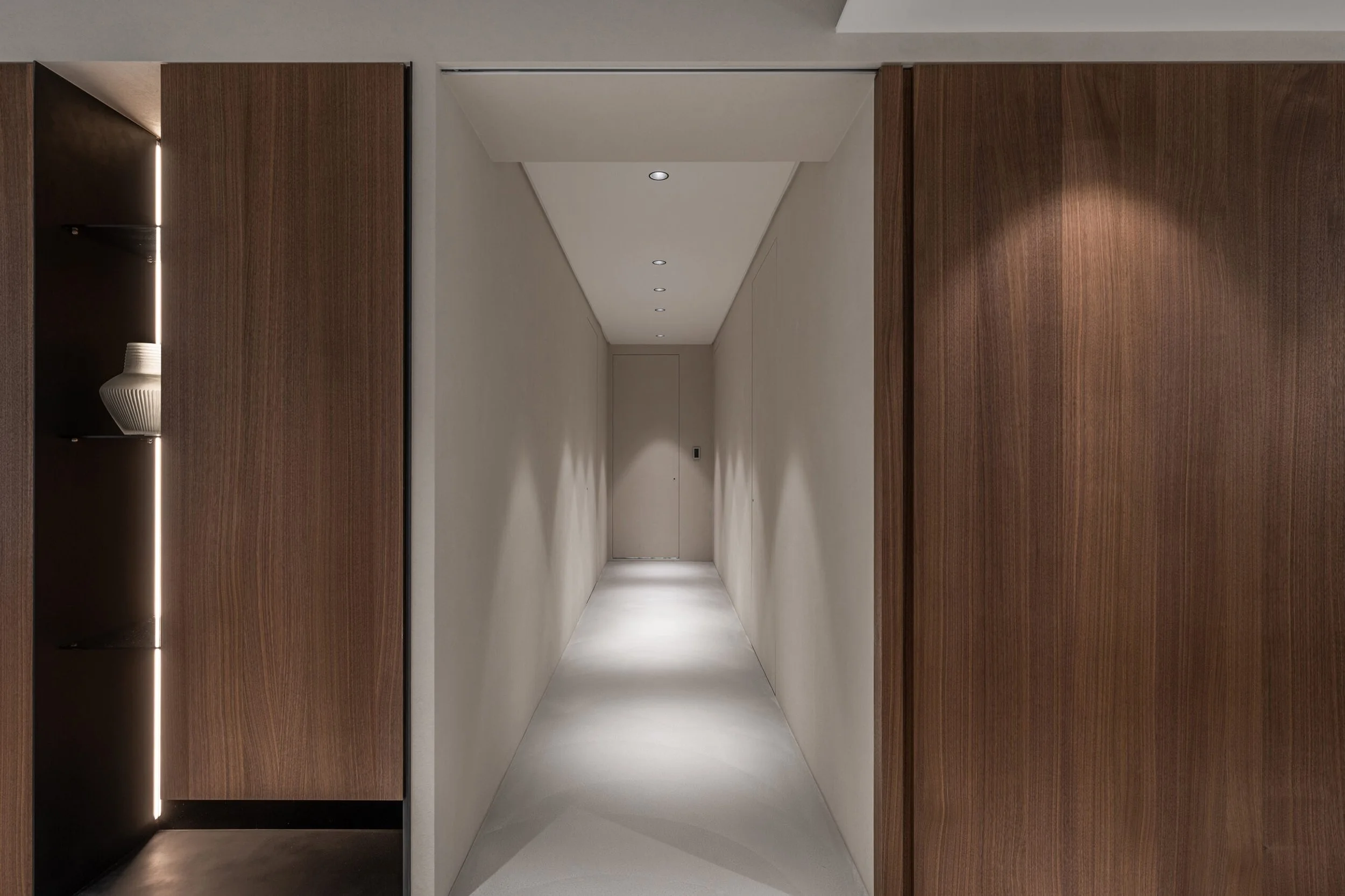 Interior hallway with white walls, ceiling, and flooring, flanked by wooden paneling on both sides, and small recessed ceiling lights.