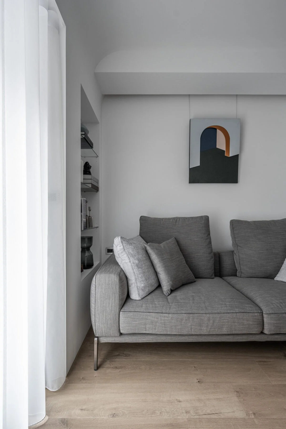 A gray sofa with multiple matching throw pillows in a minimalist living room. The wall behind has a geometric art piece with an arch and abstract shapes, with a built-in shelf on the left side with books and decorative objects.