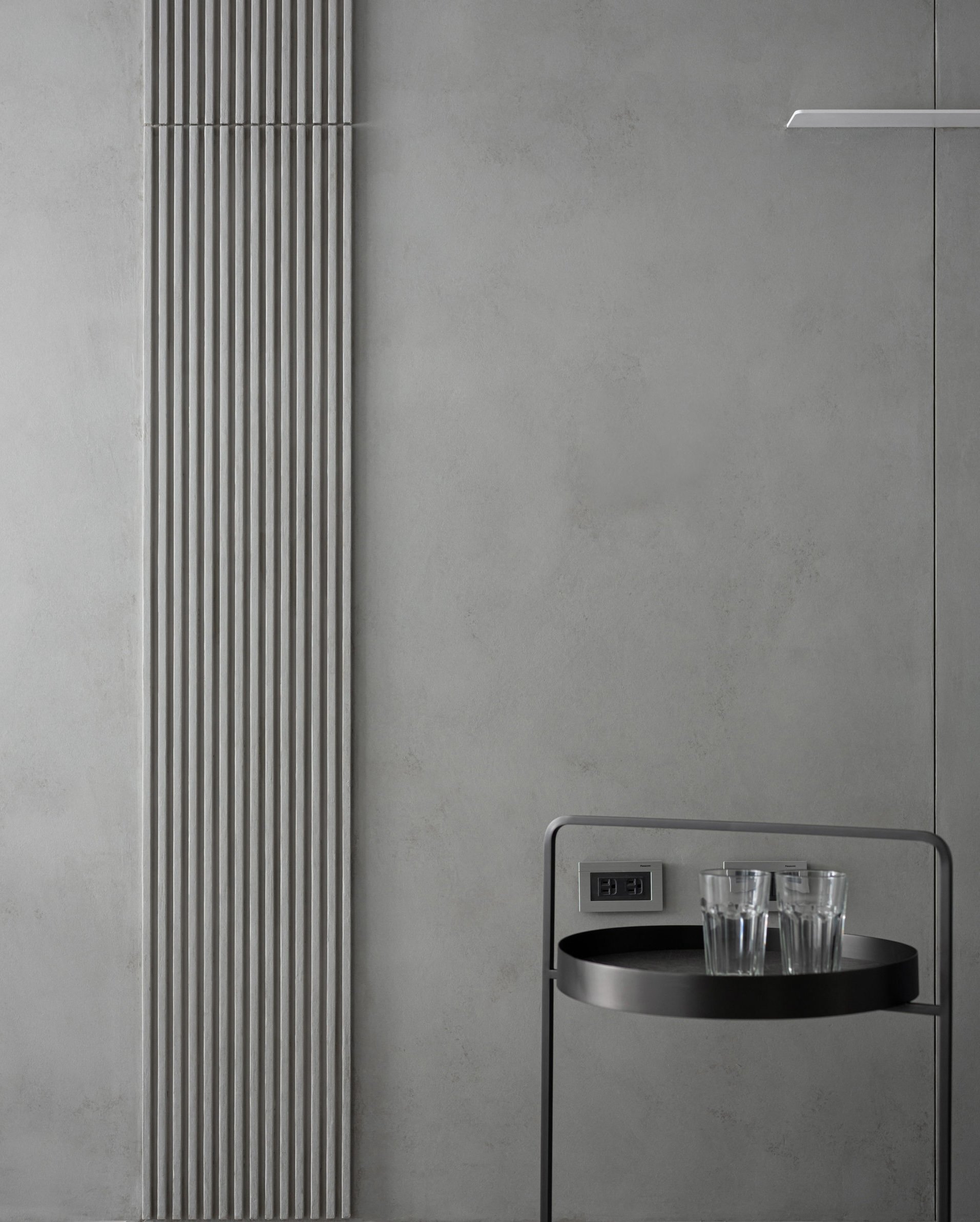 Interior wall with vertical metal radiator pipes, electrical outlet, a small stainless steel table with glasses of water underneath, and a box with a black lid.