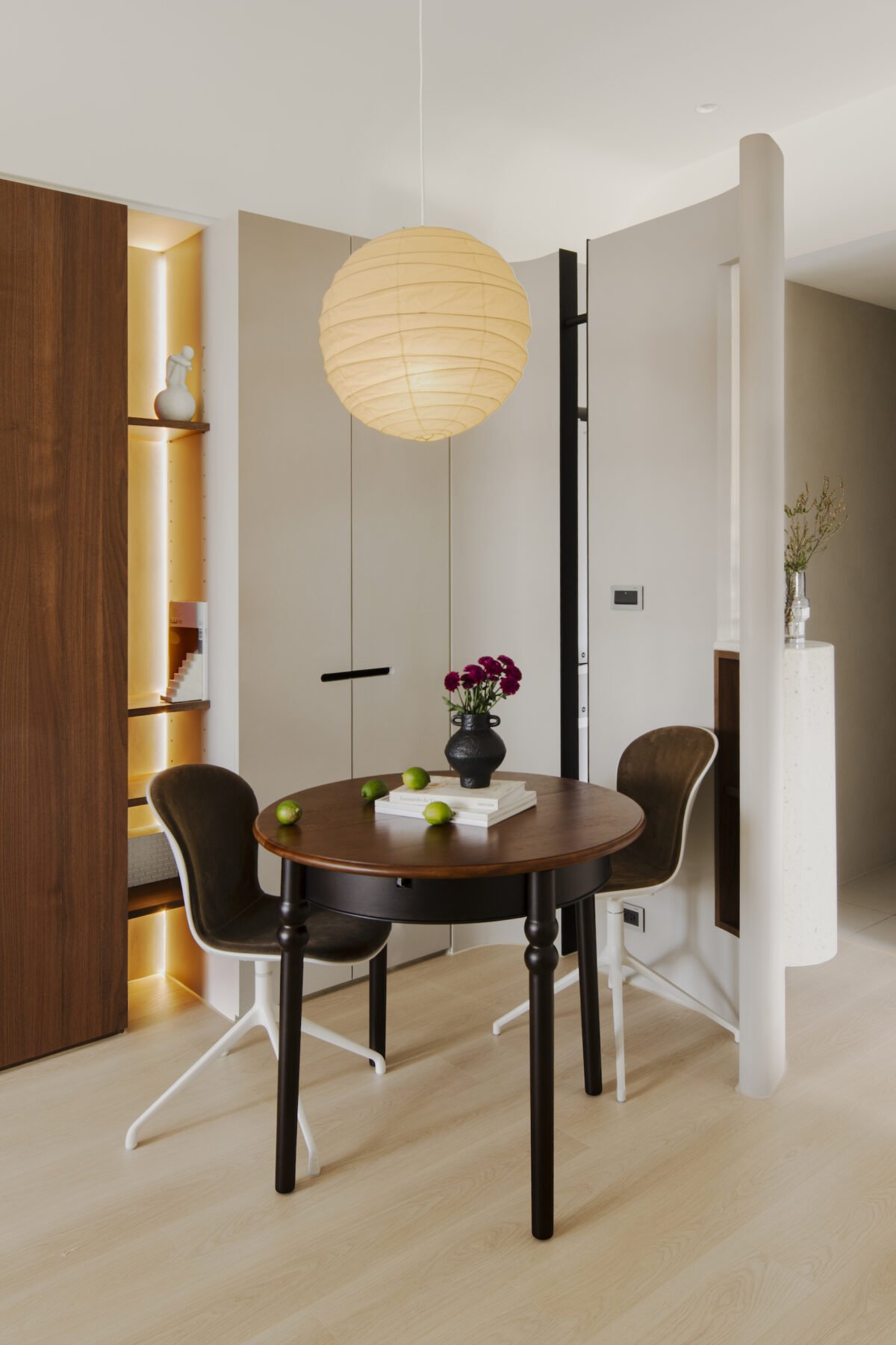 Living room with a round wooden dining table, two black chairs, a beige paper lantern ceiling light, a black vase with pink flowers, green apples, and a background with built-in shelves, a white closet door, and a small vase with flowers.