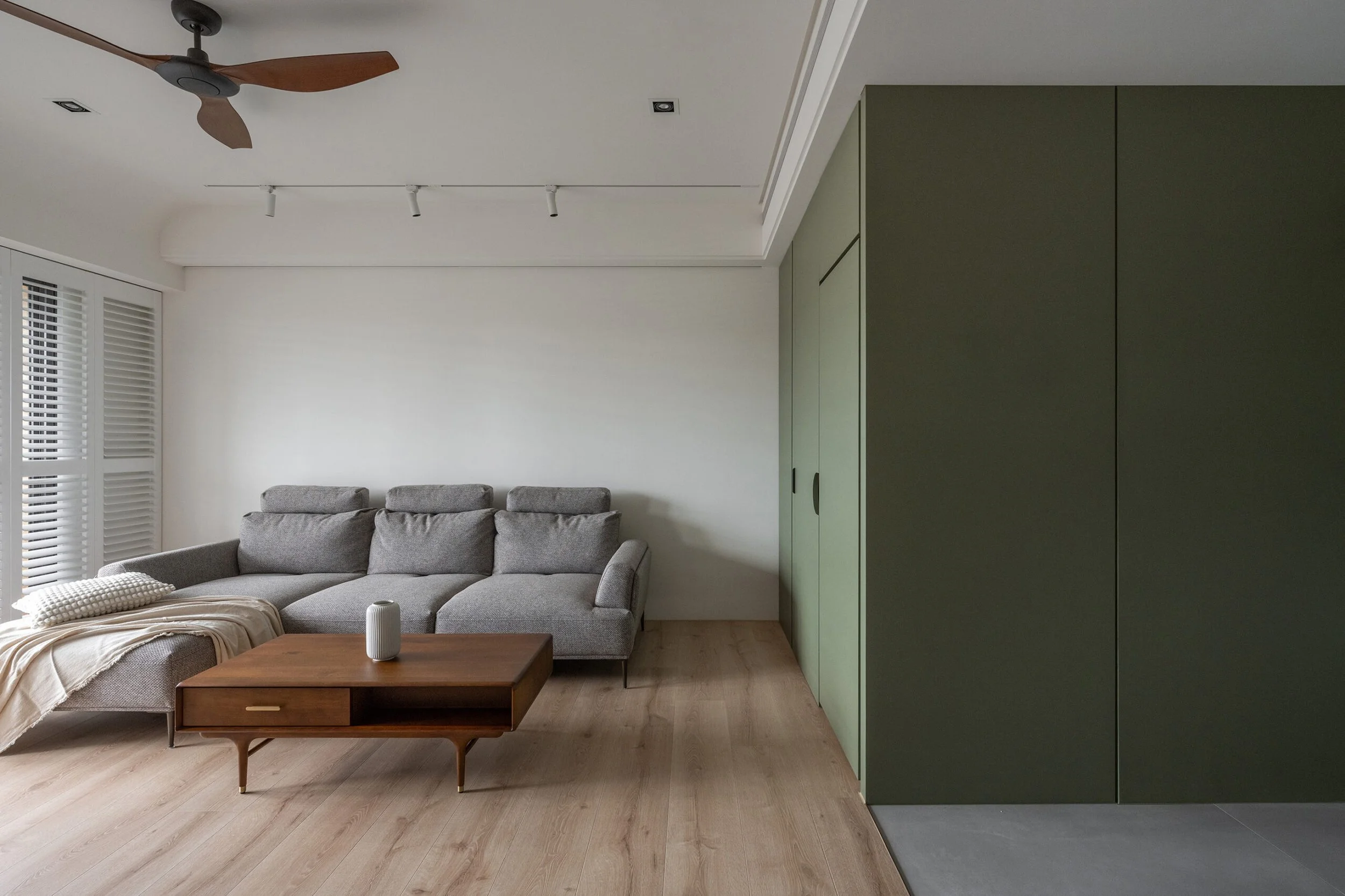 Living room with a gray sectional sofa, a wooden coffee table, green built-in cabinets, a ceiling fan with wooden blades, windows with white blinds, and a beige blanket on the sofa.