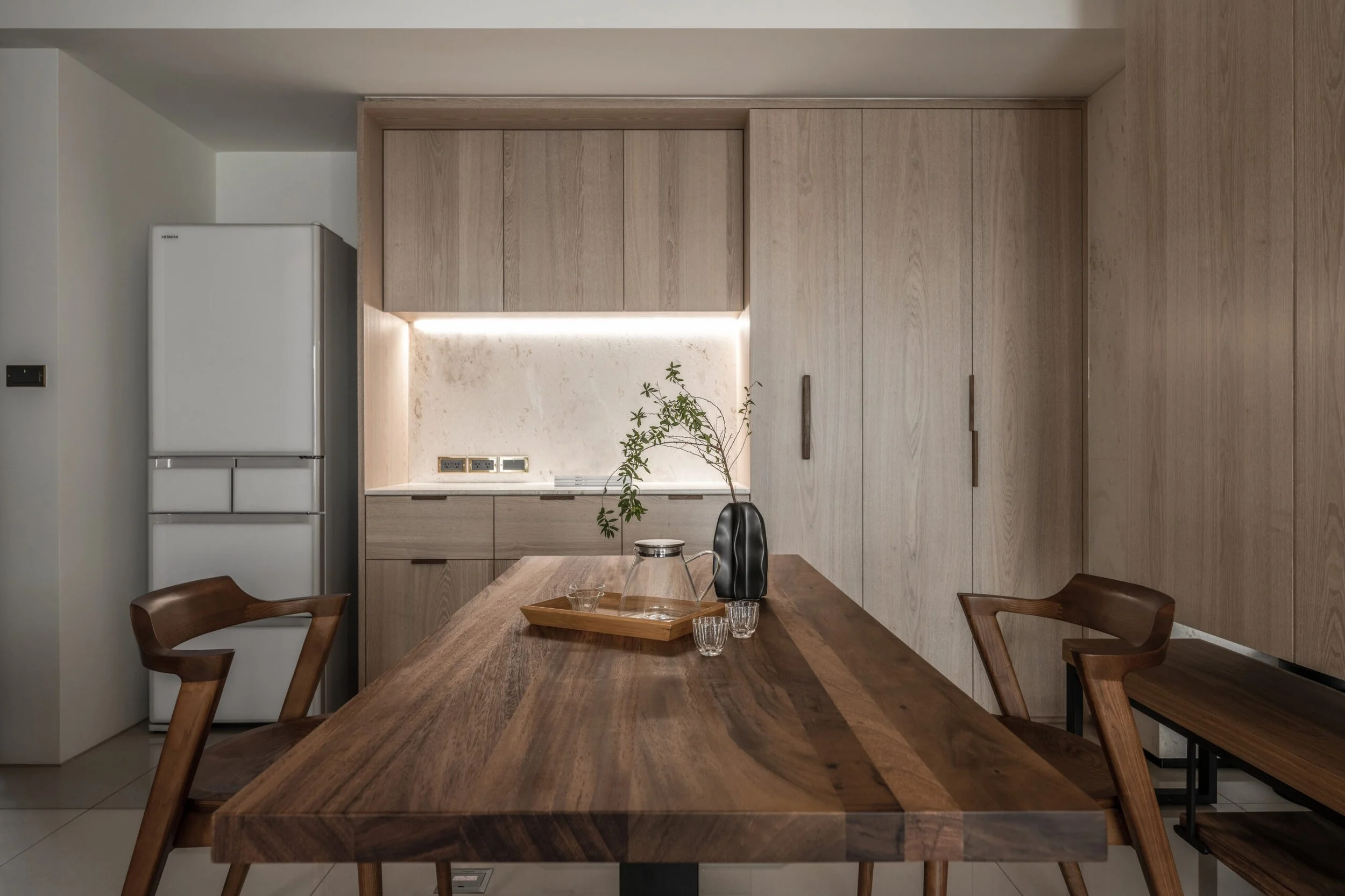 Interior of a modern kitchen with wooden cabinets, a marble backsplash, a refrigerator, and a dining table with chairs.
